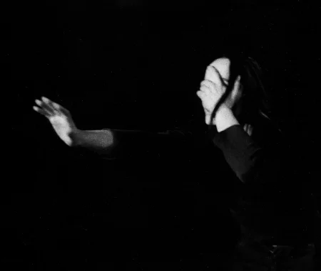 Aktorka jedną ręką zasłania twarz. Drugą rękę wyciąga przed siebie. Ma na sobie czarną koszulkę.