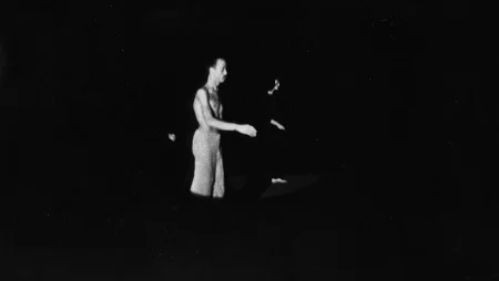 Niewyraźne zdjęcie. W świietle reflektora stoi aktor zwrócony profilem w stronę widza. Ubrany jest w jasne spodenki, ma nagi tors. Delikatnie unosi jedną rękę. Za nim stoi aktorka ubrana na czarno. Przybiera taką samą pozę jak aktor.