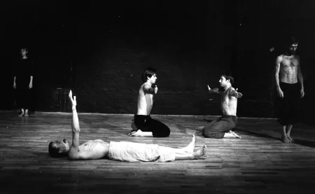 Po lewej stronie stoi zwrócona w stronę widza aktorka. Ubrana jest w czarne spodenki i czarną podkoszulkę. Jest boso. W centrum, na pierwszym planie na plecach leży aktor. Ubrany jest w jasne spodenki, ma nagi tors. Patrzy w sufit i wyciągata przed siebie jedną rękę. Za nim klęczy dwóch aktorów. Ubrani są w ciemne spodenki, mają nagie torsy. Klęczą naprzeciwko siebie i patrzą na siebie nawzajem. Mają ręce wyciągnięte na boki. Po prawej stronie stoi zwrócony w stronę widza aktor. Jest ubrany w czarne spodenki, ma nagi tors. Jest boso. Patrzy w podłogę.