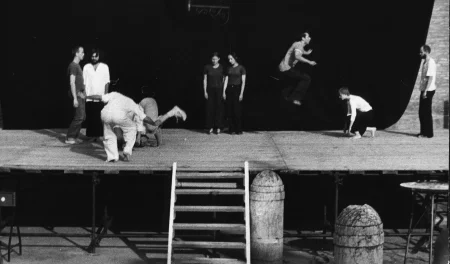 Scena plenerowa. Na scenę prowadzą drewniane schody. Z tyłu sceny ściana z czarnej płachty. Pośrodku, zza płachty wystaje latarnia. Na scenie stoją aktorzy i aktorki w różnych działaniach. Turlają się, skaczą i stoją w miejscu. Wyglądają, jakby mieli na sobie prywatne ubrania. Wspólnie tworzą choreograficzną kompozycję.