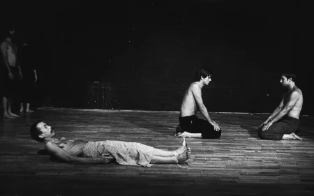 Na plecach leży aktor. Ubrany jest w jasne spodenki, ma nagi tors. Ma lekko uniesioną głowę i patrzy przed siebie. Za nim klęczy dwóch aktorów. Ubrani są w ciemne spodenki, mają nagie torsy. Klęczą naprzeciwko siebie i patrzą na siebie nawzajem. Po lewej stronie ledwo widoczne sylwetki aktorów stojących w cieniu.