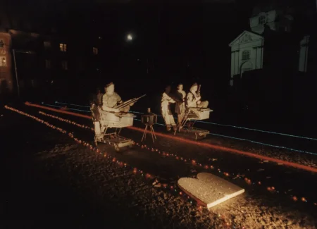 Noc. Przestrzeń miejska. Na piasku leżą trasy wyznaczone przez czerwone i niebieskie lampki. Wzdłuż dwóch tras poruszają się aktorzy i aktorki. W obu grupach jedna osoba siedzi w wózku sklepowym i trzyma przedmiot przypominający karabin. Wózki są popychane przez innych aktorów i aktorki. Wszyscy ubrani są w ubrania wojskowe. W tle widać budynki.