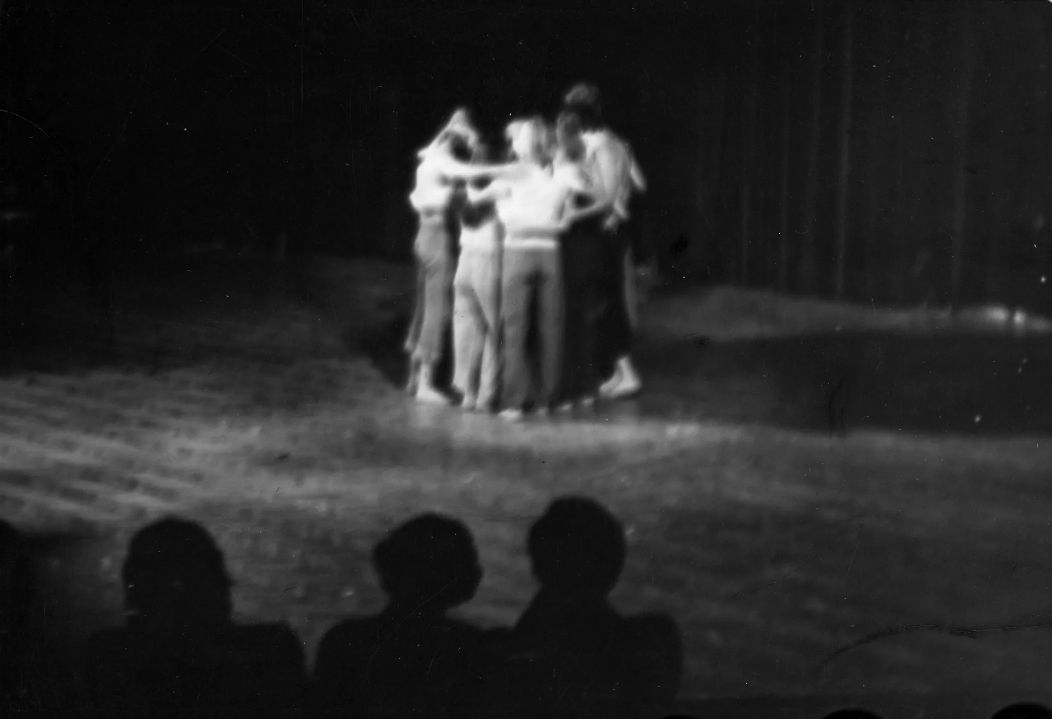 Na środku sceny w okręgu stoi grupa obejmujących się aktorów. Patrzy na nich troje widzów zwróconych do nas plecami. W tle opuszczona kurtyna.