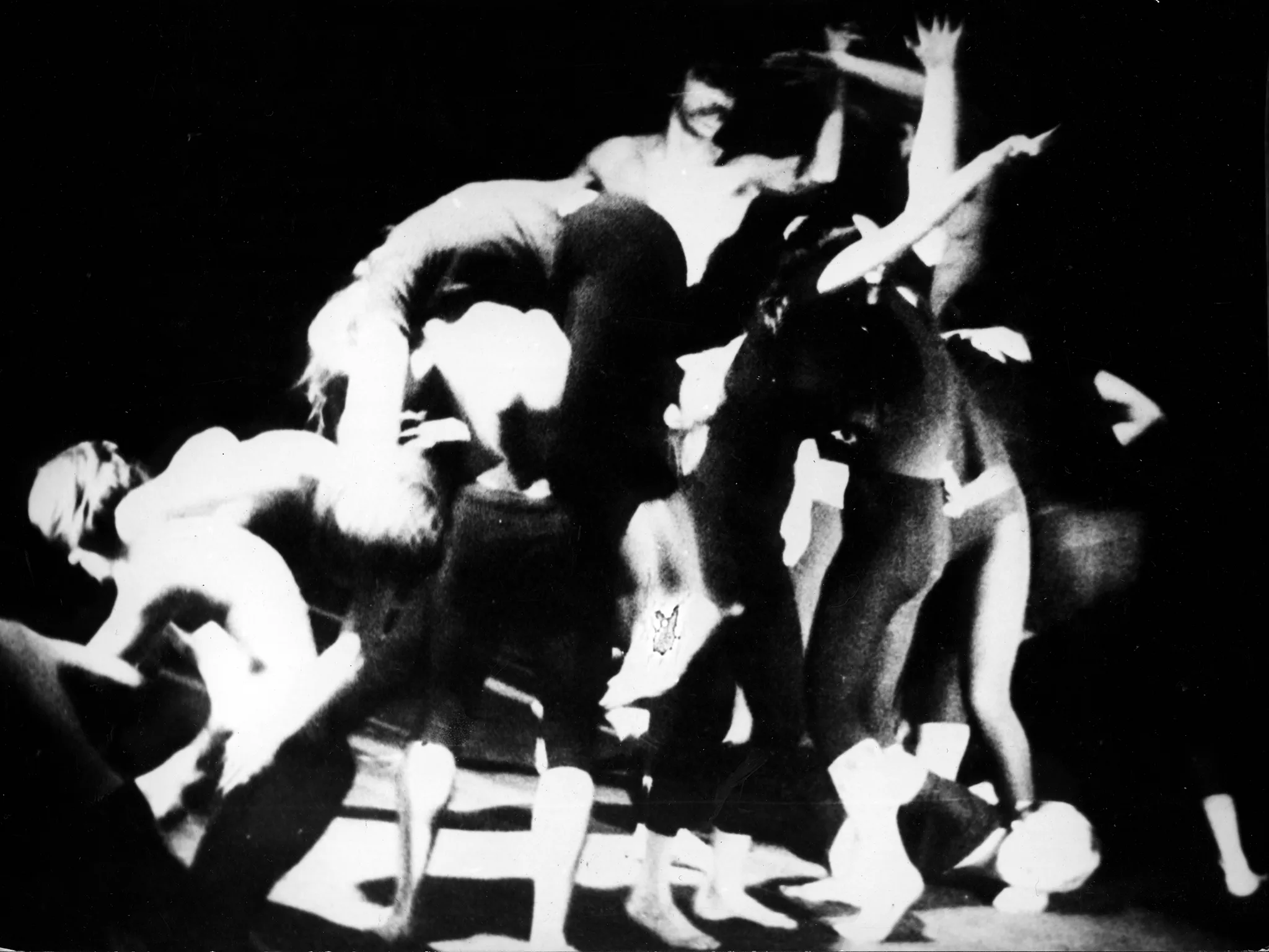 Niewyraźna grupa aktorów w akobatycznych pozach. Wszyscy są boso i stoją na czarno-białej podłodze.