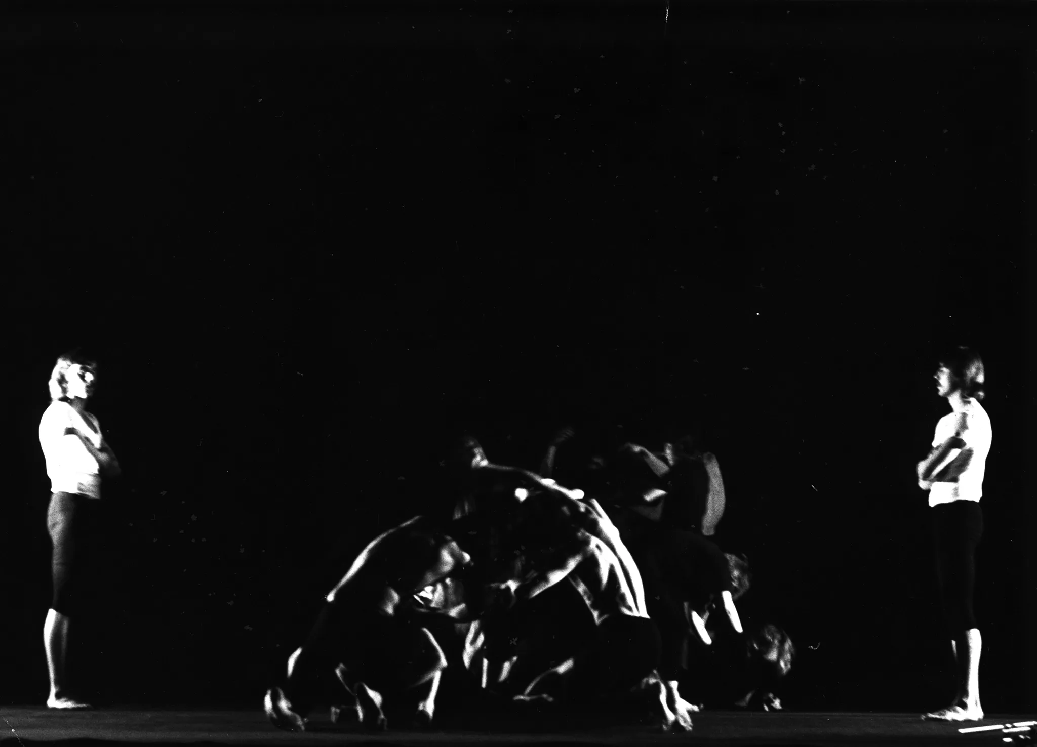 W centrum grupa aktorów w akboratycznych pozach. Na pierwszym planie dwóch wysportowanych mężczyzn bez koszulek trzyma się wzajemnie tworząc okrąg. Po lewej i prawej stronie stoi jeden aktor w białym podkoszulku i czarnych spodenkach. Obaj patrzą grupę aktorów w centrum.