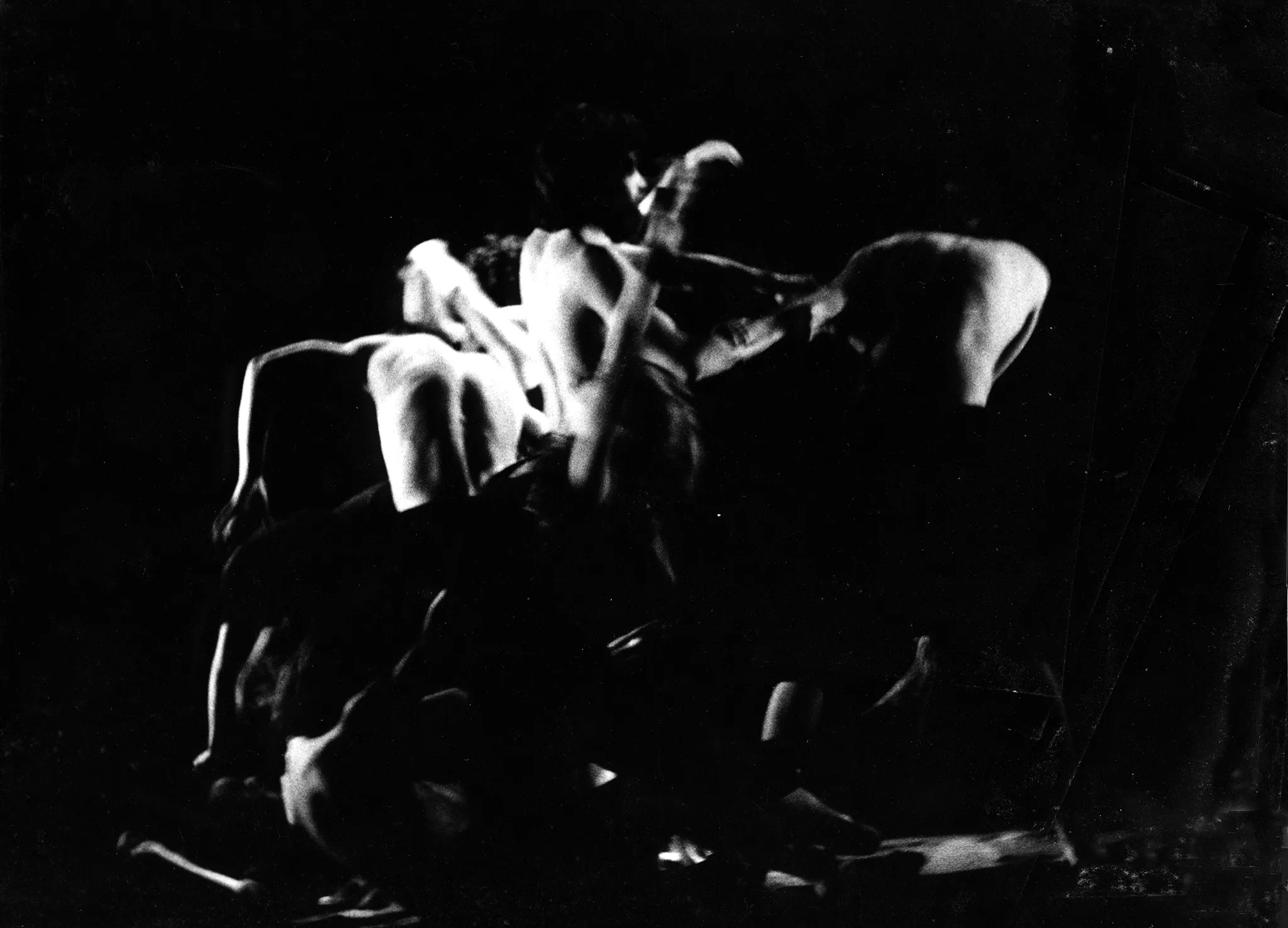 Niewyraźna grupa aktorów bez koszulek w akrobatycznych pozach. Po lewej stronie na podłodze leży kość.