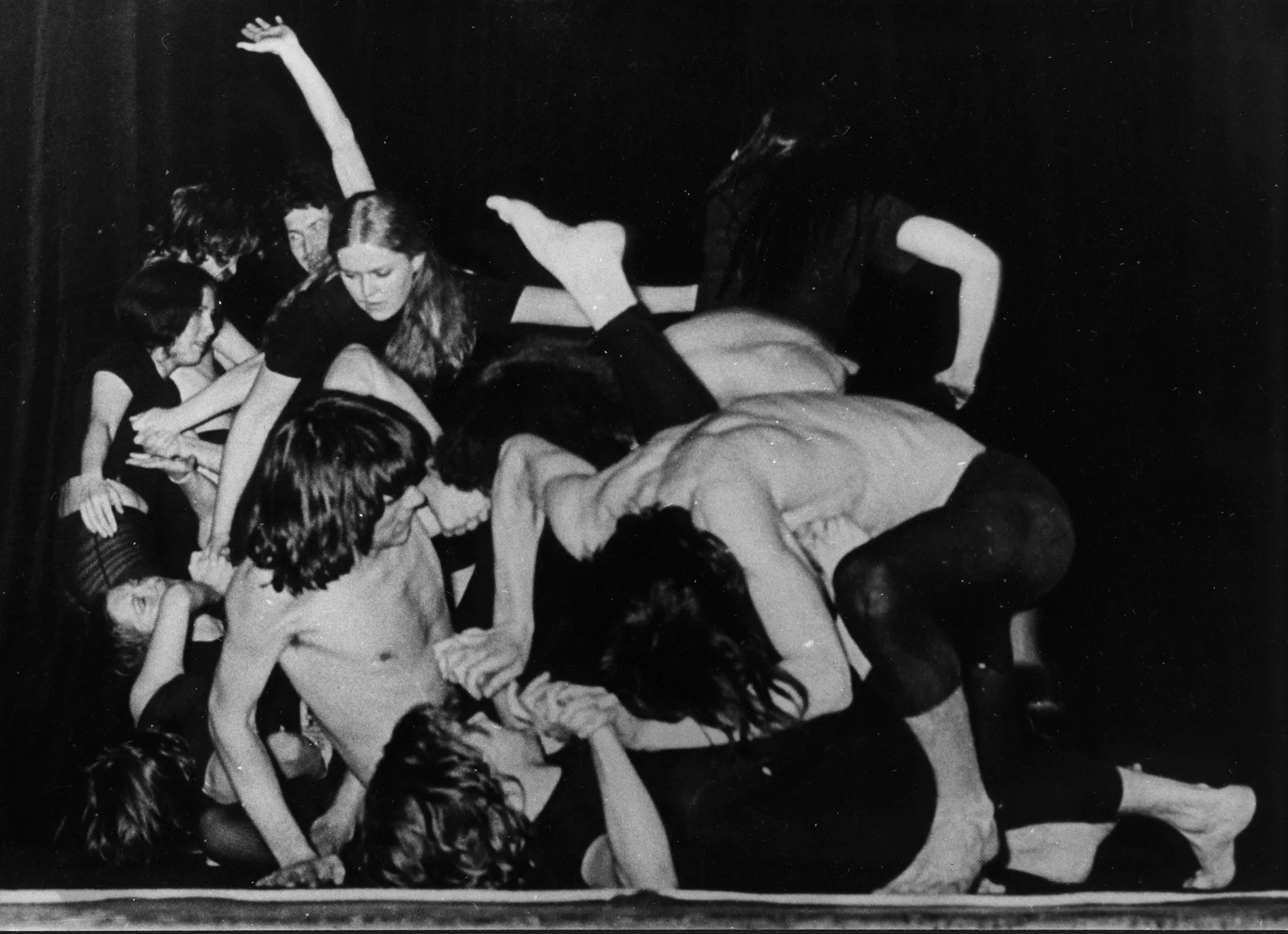 Skłębiona grupa aktorów i aktorek. Niektórzy leżą na podłodze, inni leżą na nich, tworząc kolejne warstwy. Ich ciała zwrócone są w różnych kierunkach, tworząc wrażenie chaosu.