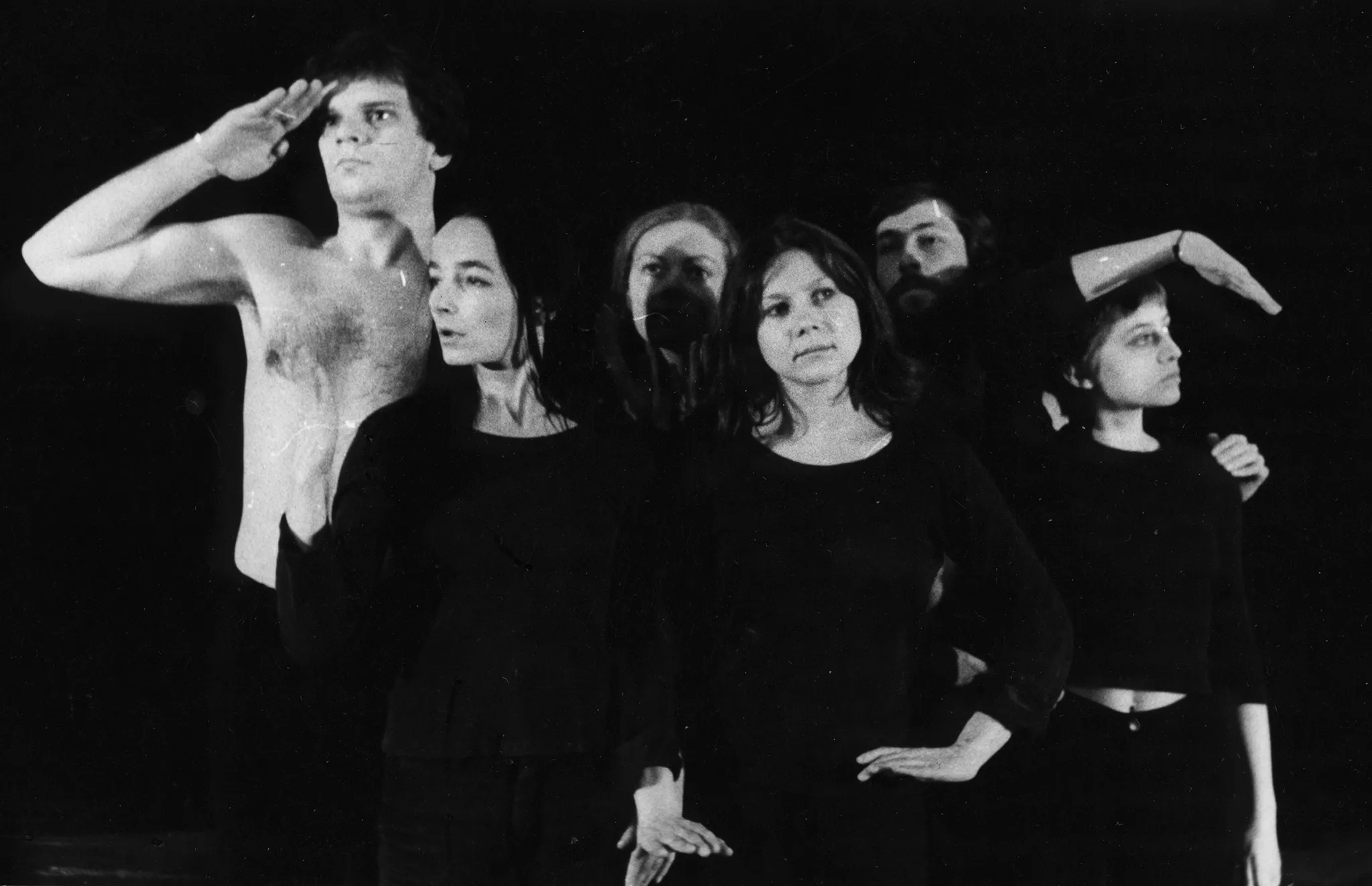 Dwóch aktorów i cztery aktorki. Aktor stojący po lewej stronie nie ma koszulki, jest poważny i trzyma dłoń przy swoim czole. Aktorki ubrane są na czarno i wykonują gesty dłońmi. Stojący za nimi aktor jest ledwo widoczny, ale widać, że patrzy przed siebie.