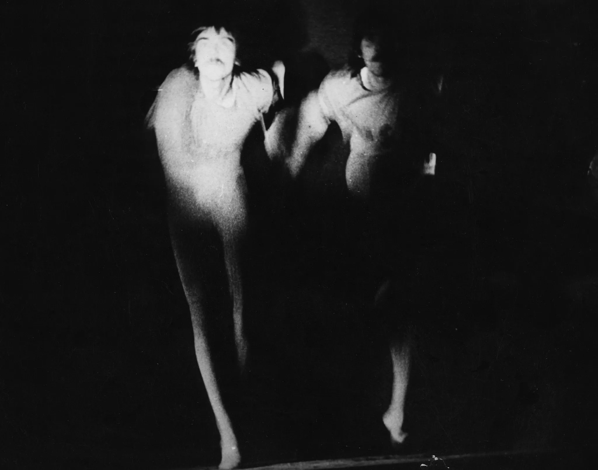 Niewyraźne, rozmazane zdjęcie. Dwóch aktorów zwróconych w naszą stronę trzyma się za ręce i biegnie przed siebie. Na twarz aktora po lewej stronie pada światło. Twarz aktora po prawej stronie jest ukryta w cieniu.