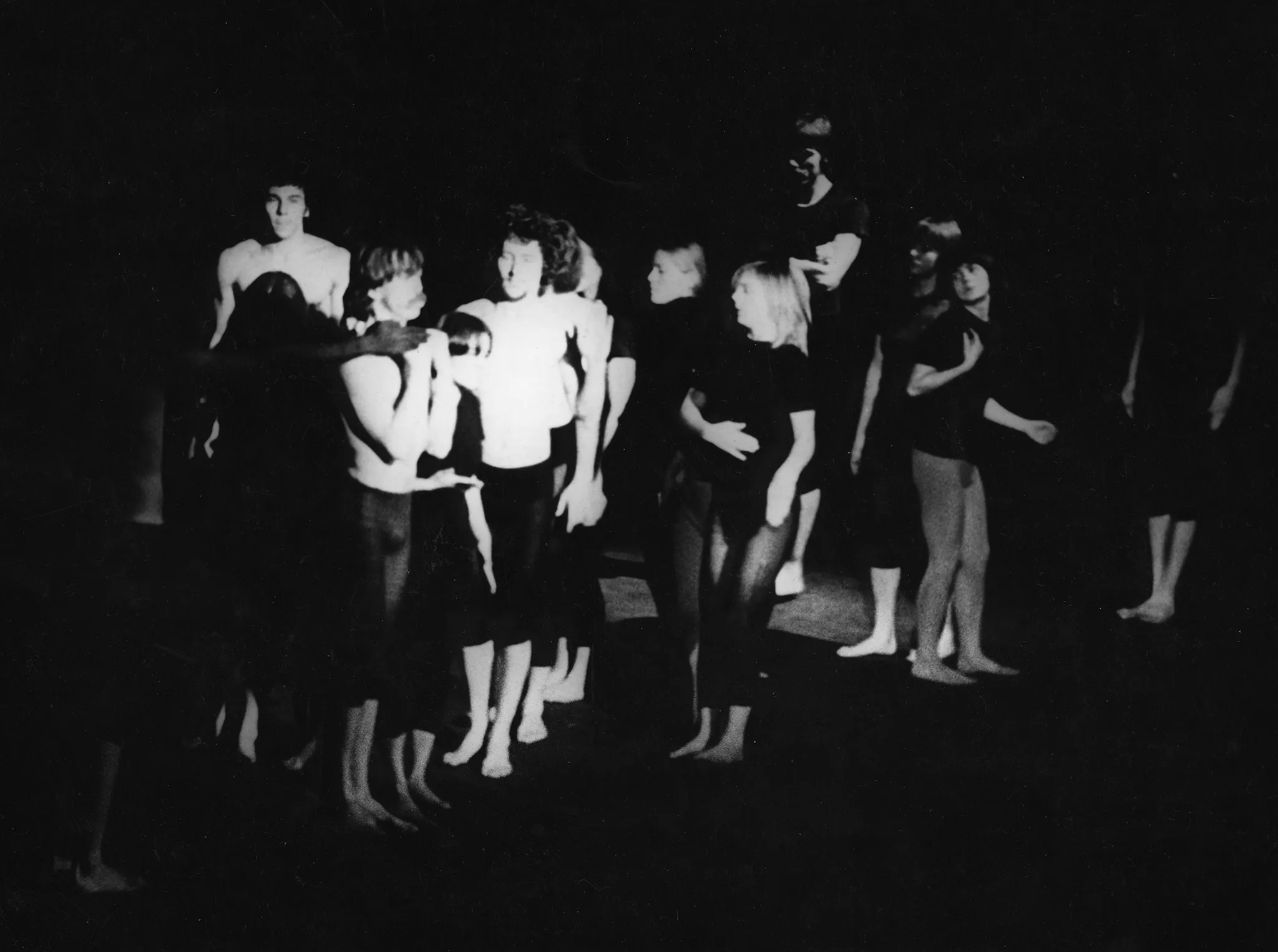 Grupa aktorów i aktorek. Stojący po lewej stronie ubrani są w czarne spodenki, nie mają na sobie koszulek. Jeden z aktorów trzyma na plecach czarną lalkę przypominającą człowieka. Stojący po prawej stronie ubrani są w czarne spodenki i czarne podkoszulki. Jedna z aktorek nie ma na sobie spodenek, prawdopodobnie jest naga od pasa w dół.