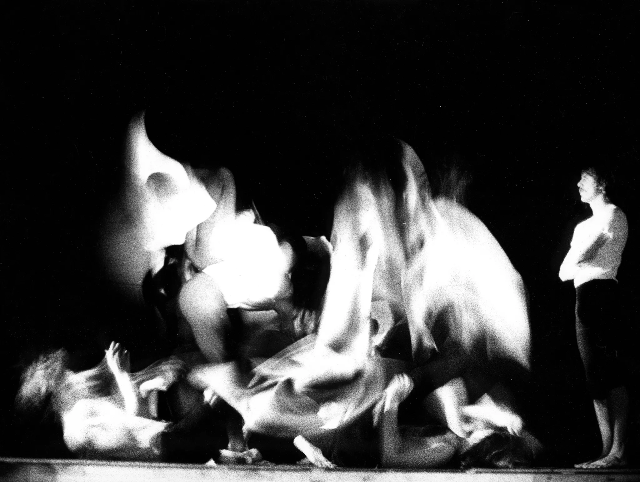 Niewyraźne, rozmazane zdjęcie. Grupa aktorów skacze i leży na pustej scenie. Po prawej stronie stoi aktor ubrany w biały podkoszulek i czarne spodenki. Jest bosy. Patrzy na grupę aktorów nie wyrażając emocji.