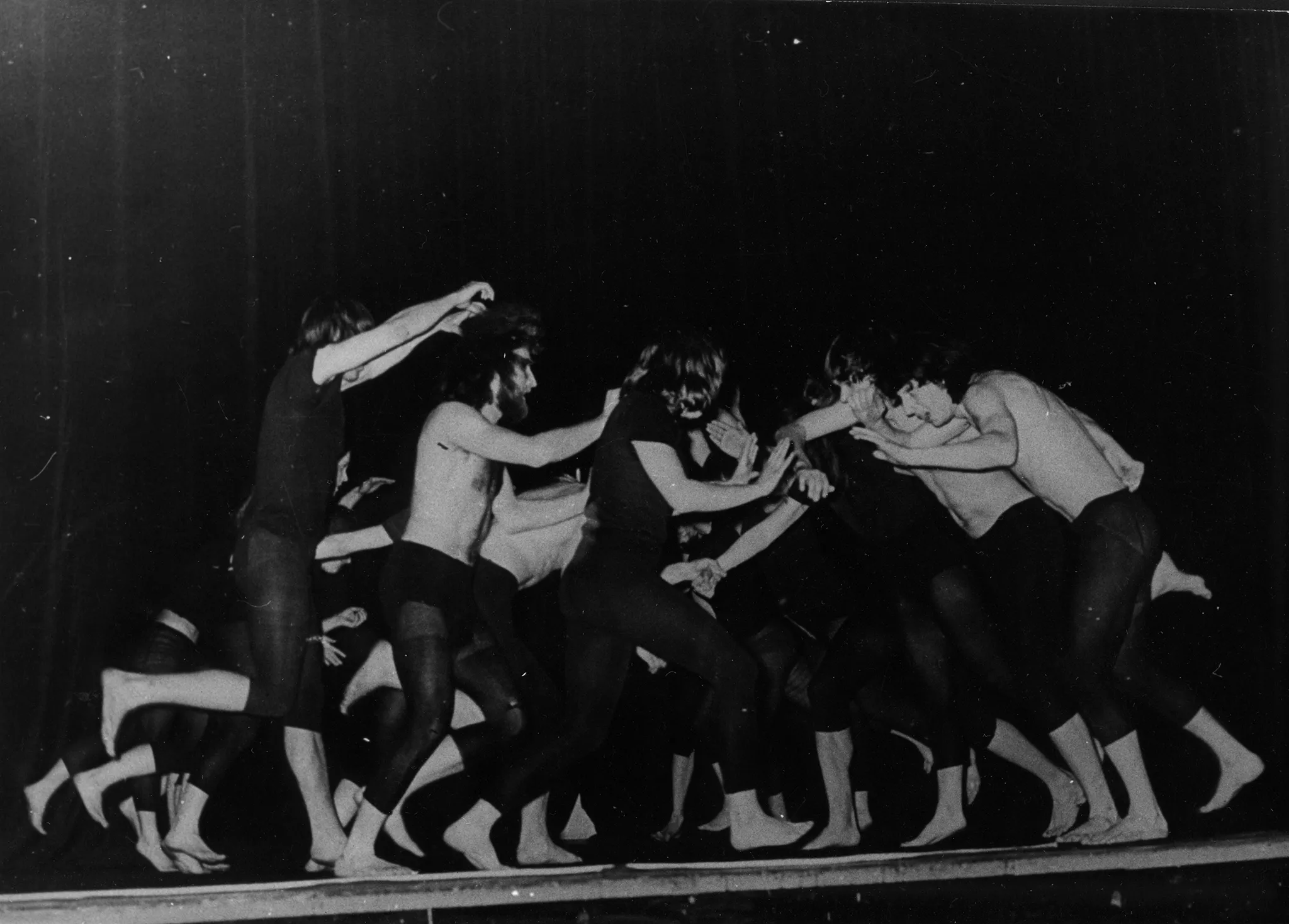Na pustej scenie kłębi się grupa aktorów. Wszyscy są boso. Niektórzy ubrani są w czarne spodenki i czarne podkoszulki, inni mają nagi tors. Ich zbiorowy ruch sprawia wrażenie chaosu. Z tyłu widać lekko oświetloną opuszczoną kurtynę.