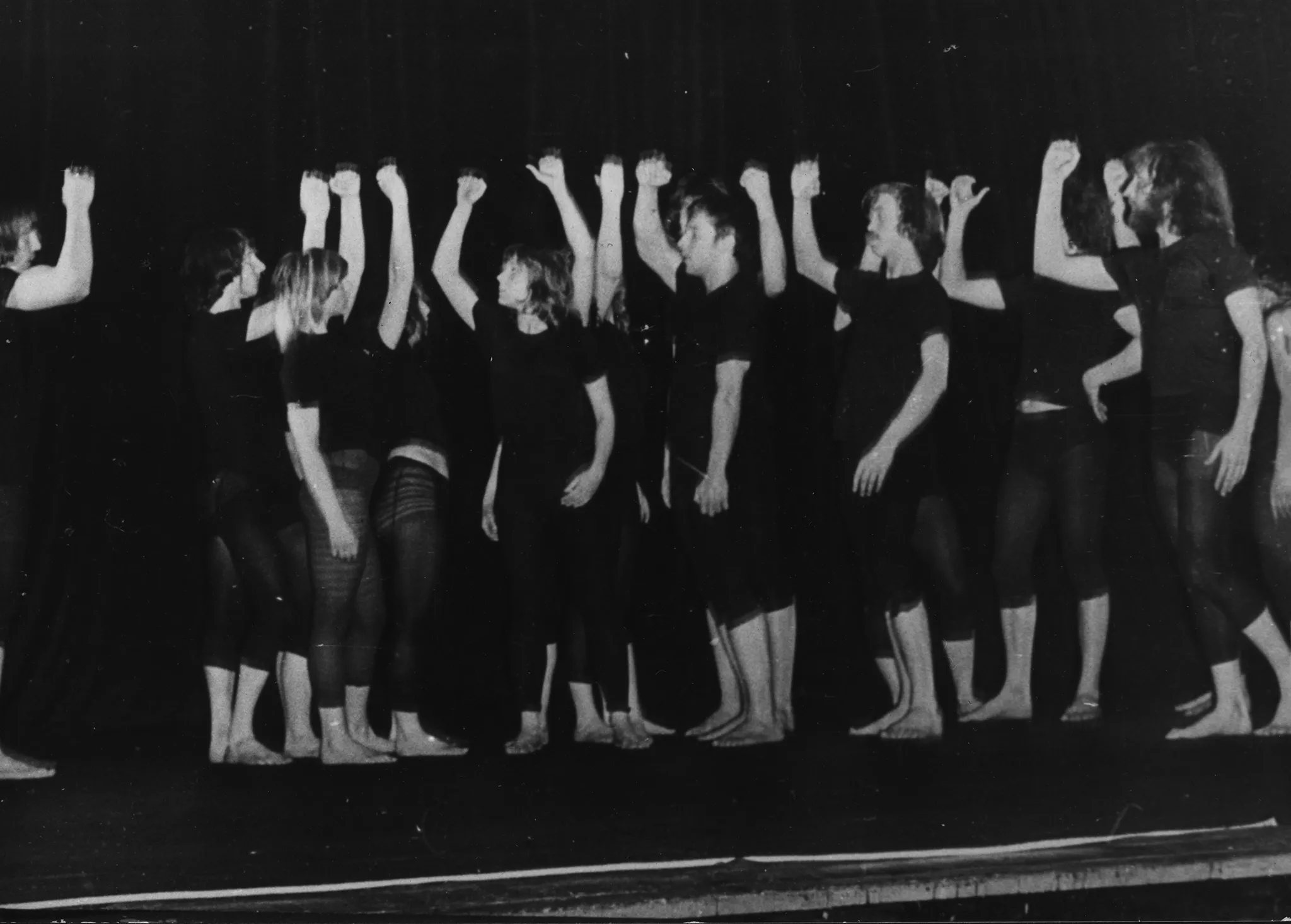 Grupa aktorów i aktorek stoi boso na pustej scenie. Są ubrani w czarne podkoszulki i czarne spodenki, u niektórych prześwitujące. Każdy trzyma w górze jedną lekko zgiętą rękę. Stojący po lewej stronie patrzą w prawo, a stojący po stronie prawej w lewo.