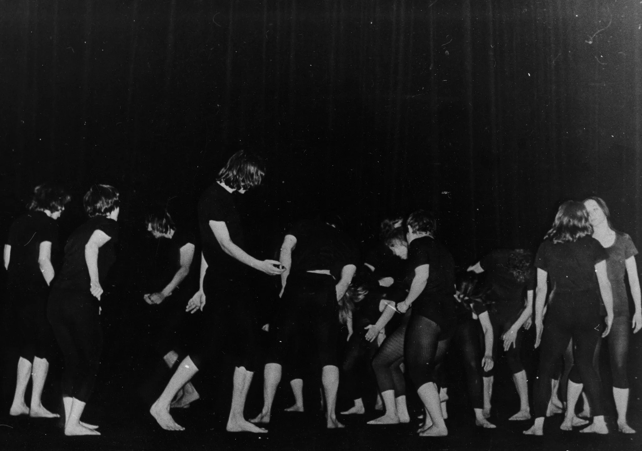 Grupa aktorów i aktorek stoi boso na pustej scenie. Są ubrani w czarne podkoszulki i czarne spodenki, u niektórych prześwitujące. Każdy trzyma w górze jedną lekko zgiętą rękę. Stojący po lewej stronie patrzą w prawo, a stojący po stronie prawej w lewo.