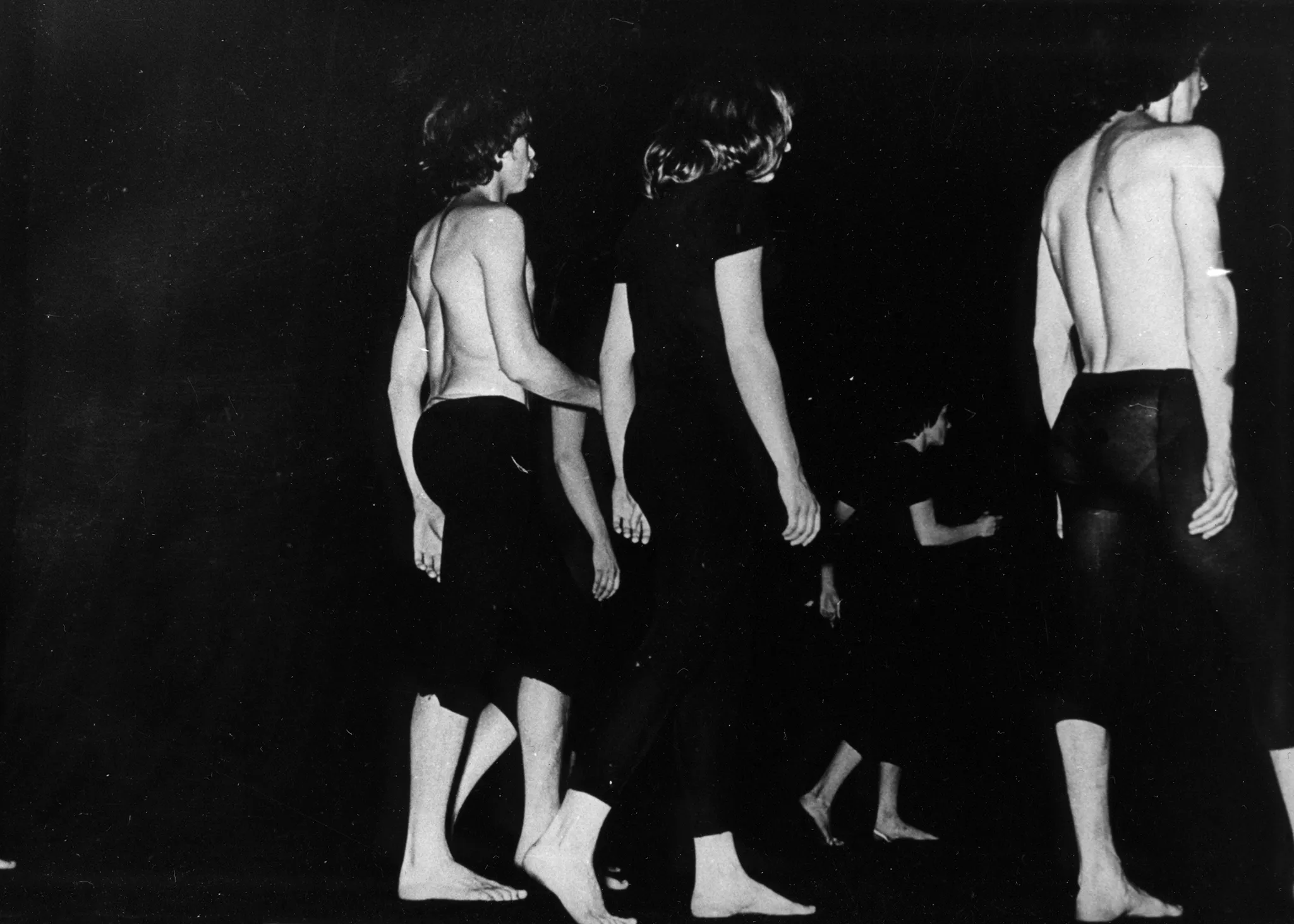 Grupa bosych aktorów idzie w prawą stronę. Aktorka w centrum ubrana jest w czarną koszulkę i czarne spodenki. Ma długie włosy. Po obu jej stronach idą aktorzy w czarnych spodenkach, bez koszulek. W oddali średnio widoczny aktor ubrany tak samo jak aktorka.