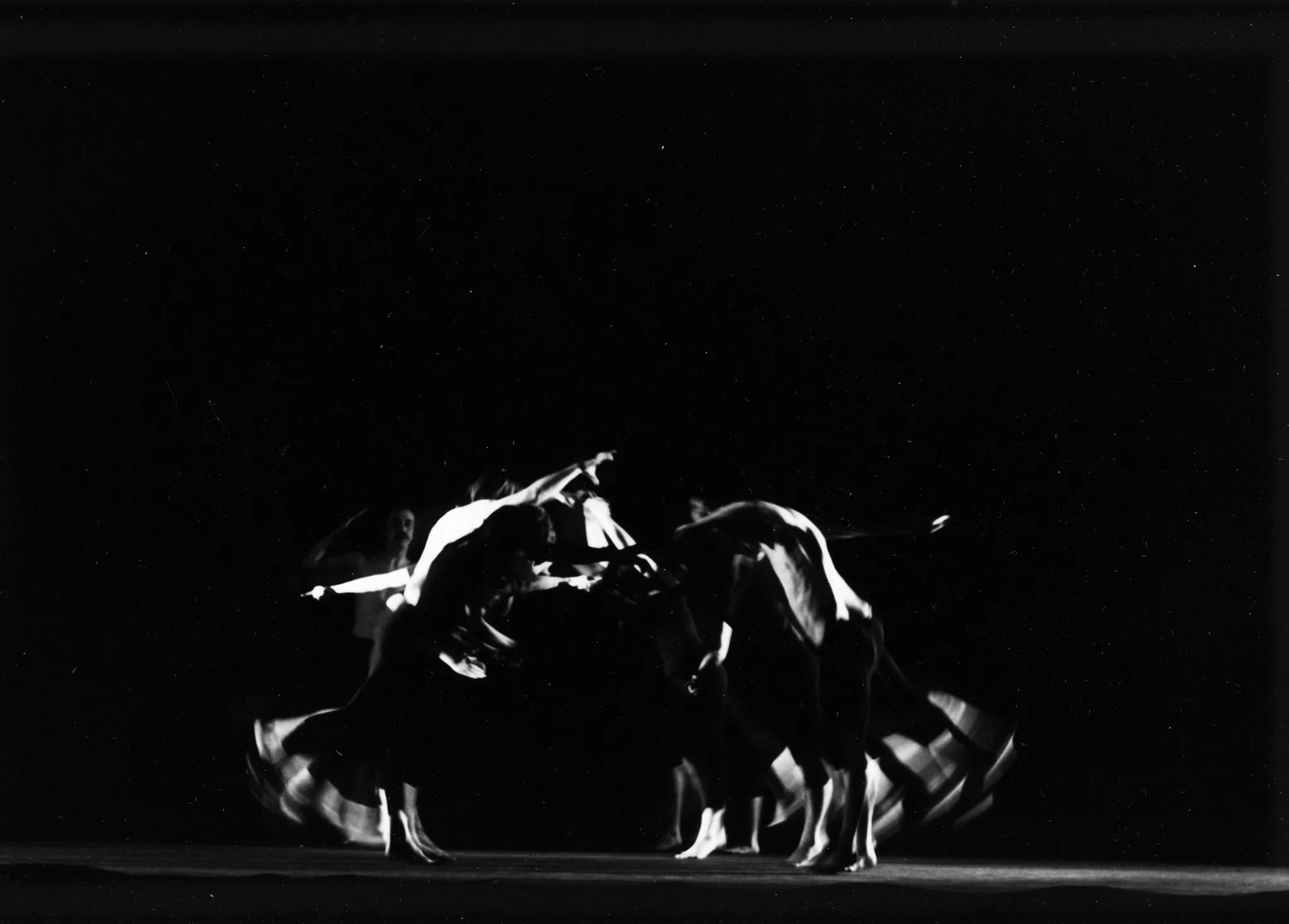 Po lewej i prawej stronie pustej sceny stoją skłonieni aktorzy zwróceni do siebie twarzami. Są boso, mają czarne krótkie spodenki, niektórzy nie mają koszulek. Wyglądają, jakby chcieli iść w stronę aktorów z drugiej strony sceny. Za grupą aktorów samotnie stoi aktor bez koszulki z wyciągniętą do góry ręką. Patrzy na grupę.