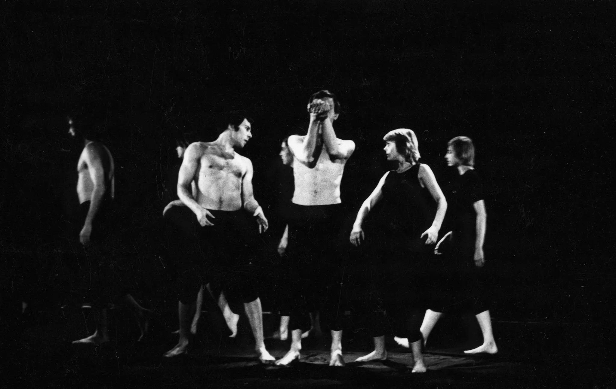 W centrum pustej sceny stoi bosy aktor ubrany w czarne spodenki, nie ma koszulki. Zasłania twarz rękami. Po jego lewej stronie stoi aktor ubrany tak samo. Po prawej stronie stoi aktorka ubrana w czarne spodenki i czarną koszulkę. Aktor i aktorka patrzą na siebie. Za ich plecami w rządku w lewą stronę idą aktorzy i aktorki. Niektórzy są ubrani w czarne koszulki, inni nie mają koszulek.