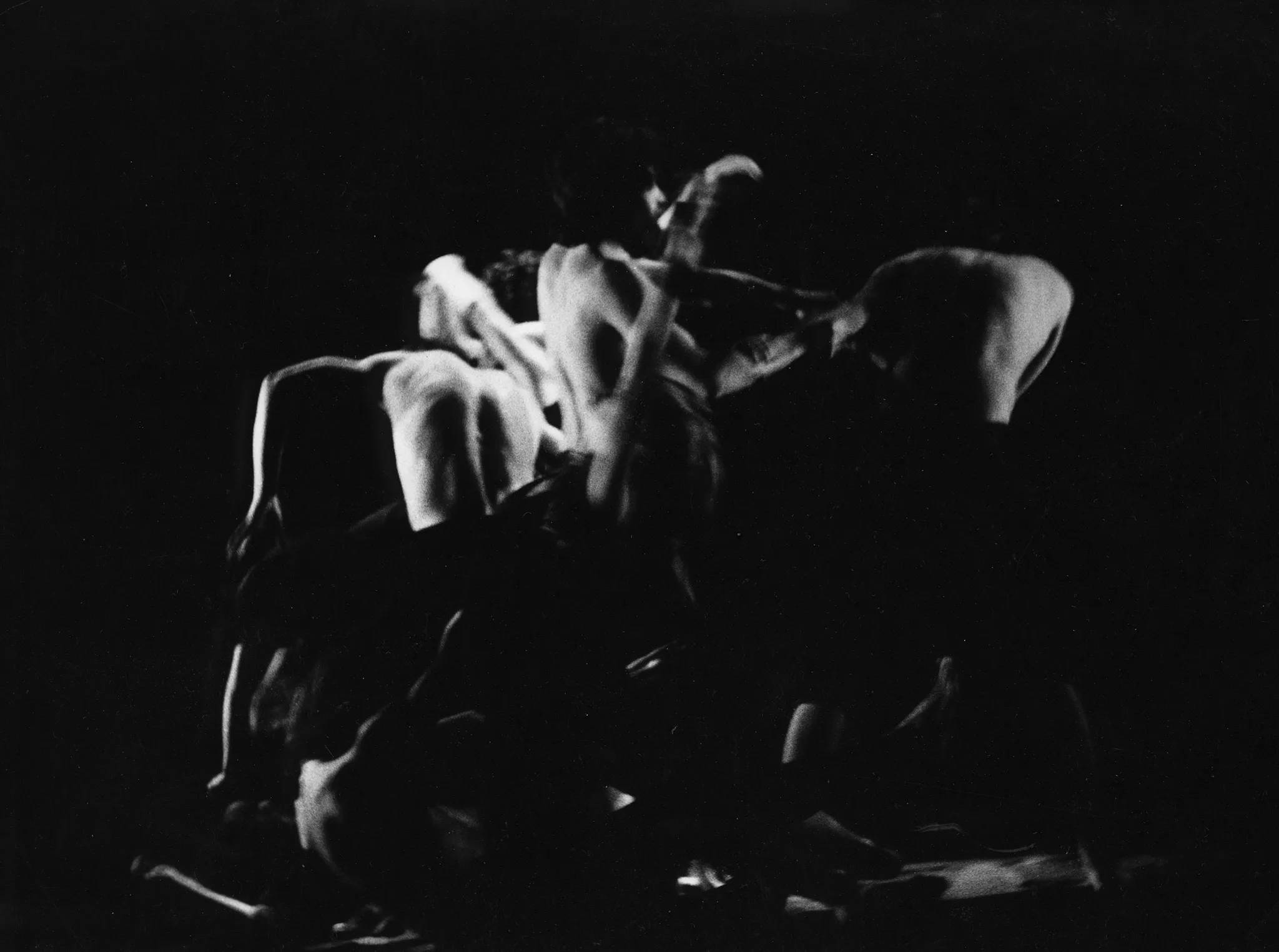 Niewyraźna grupa aktorów bez koszulek w akrobatycznych pozach. Po lewej stronie na podłodze leży kość.