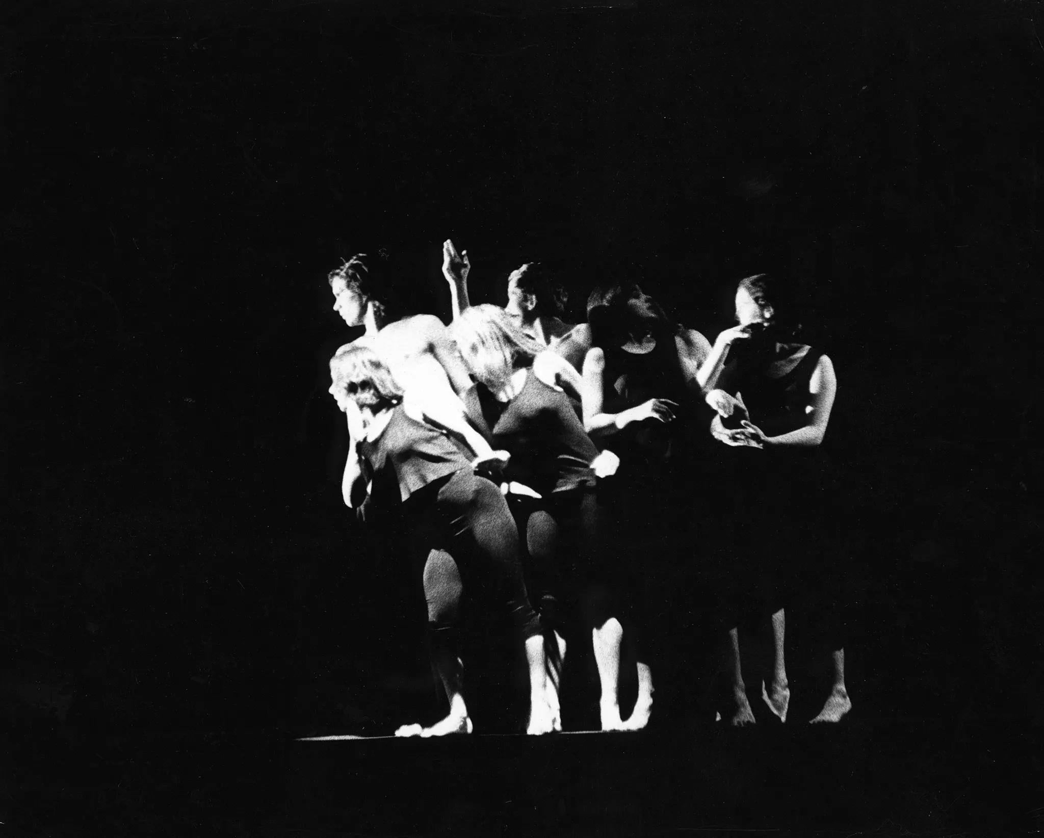 Ściśnięta grupa aktorów i aktorek stoi na pustej scenie. Są boso, ubrani w czarne spodenki i czarne podkoszulki. Niektórzy nie mają podkoszulek. Aktorzy i aktorki po lewej stronie z zaciekawieniem patrzą w lewą stronę. Dwie aktorki po prawej stronie są odrębne od grupy. Są ze sobą w interakcji.
