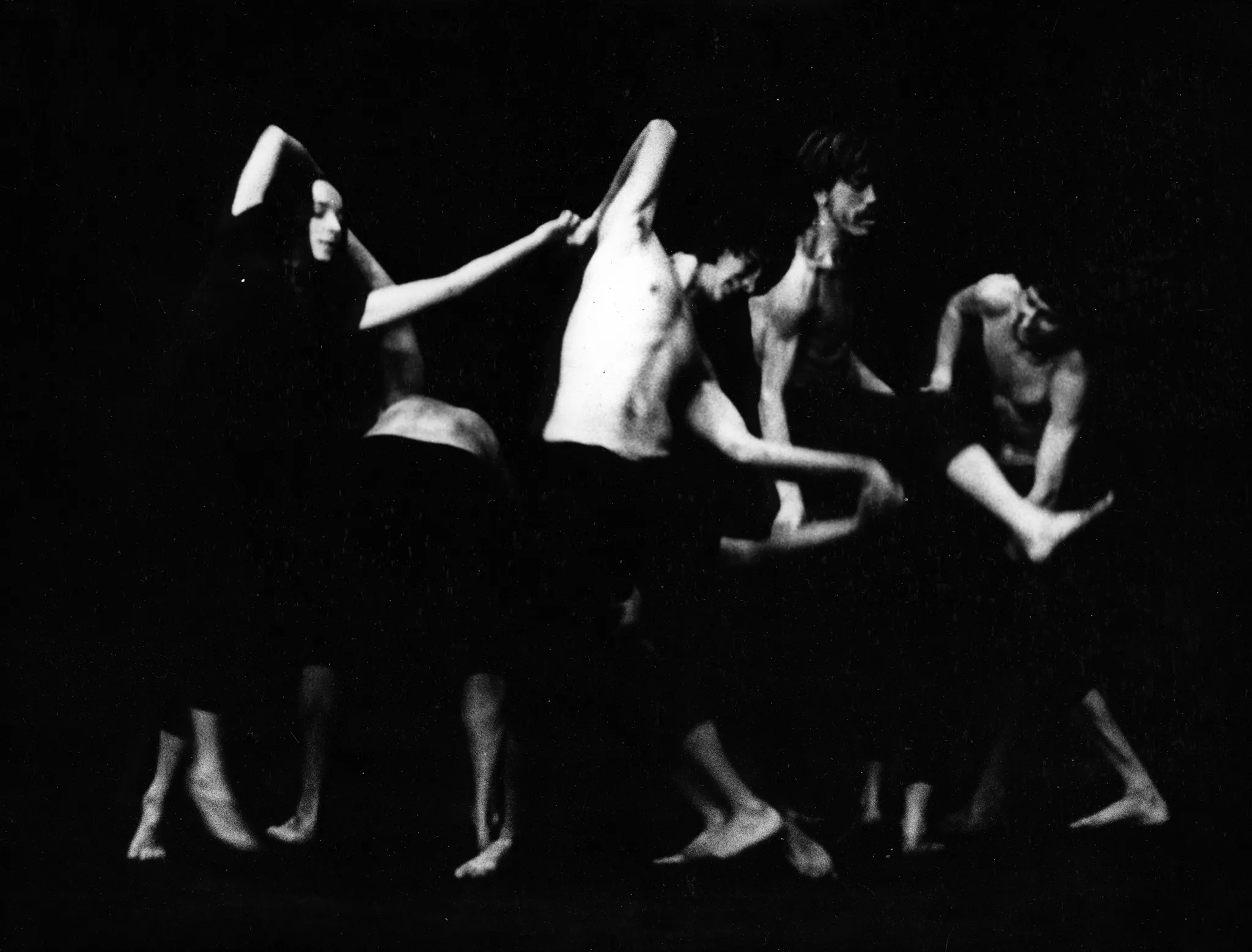 Aktorka i grupa aktorów stoją w szeregu. Trzymają się za ręce. Ich ciała są wygięte w różne strony. Aktorka jest ubrana w czarne spodenki i czarną podkoszulkę. Aktorzy nie mają podkoszulek. Wszyscy są boso.