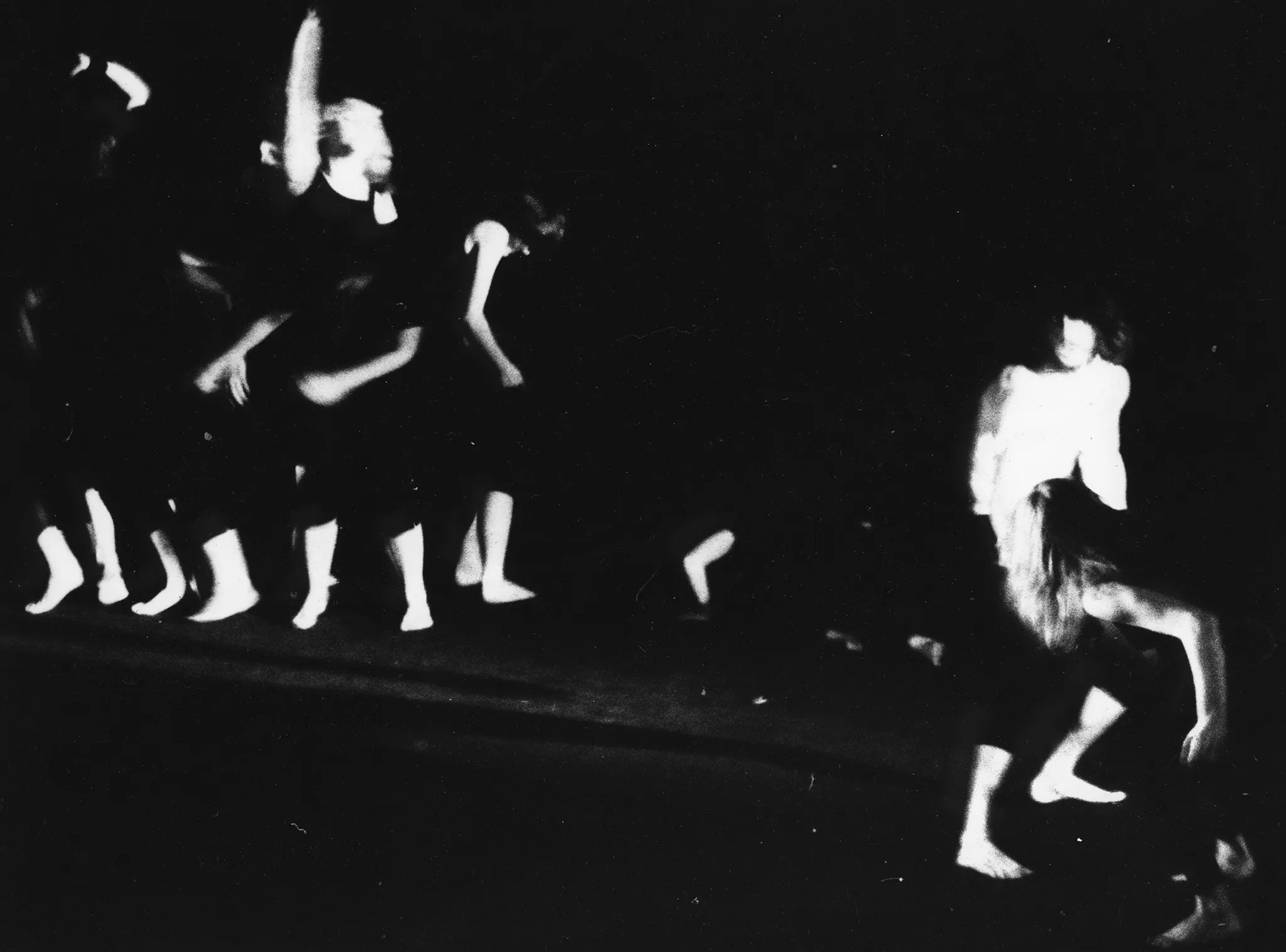 Niewyraźne, rozmazane zdjęcie. Po lewej stronie bosa grupa aktorów ubranych w czarne spodenki i czarne podkoszulki wygląda jakby tańczyła. Po prawej stronie dwie osoby odłączone od grupy stoją w ekspresyjnych pozach.