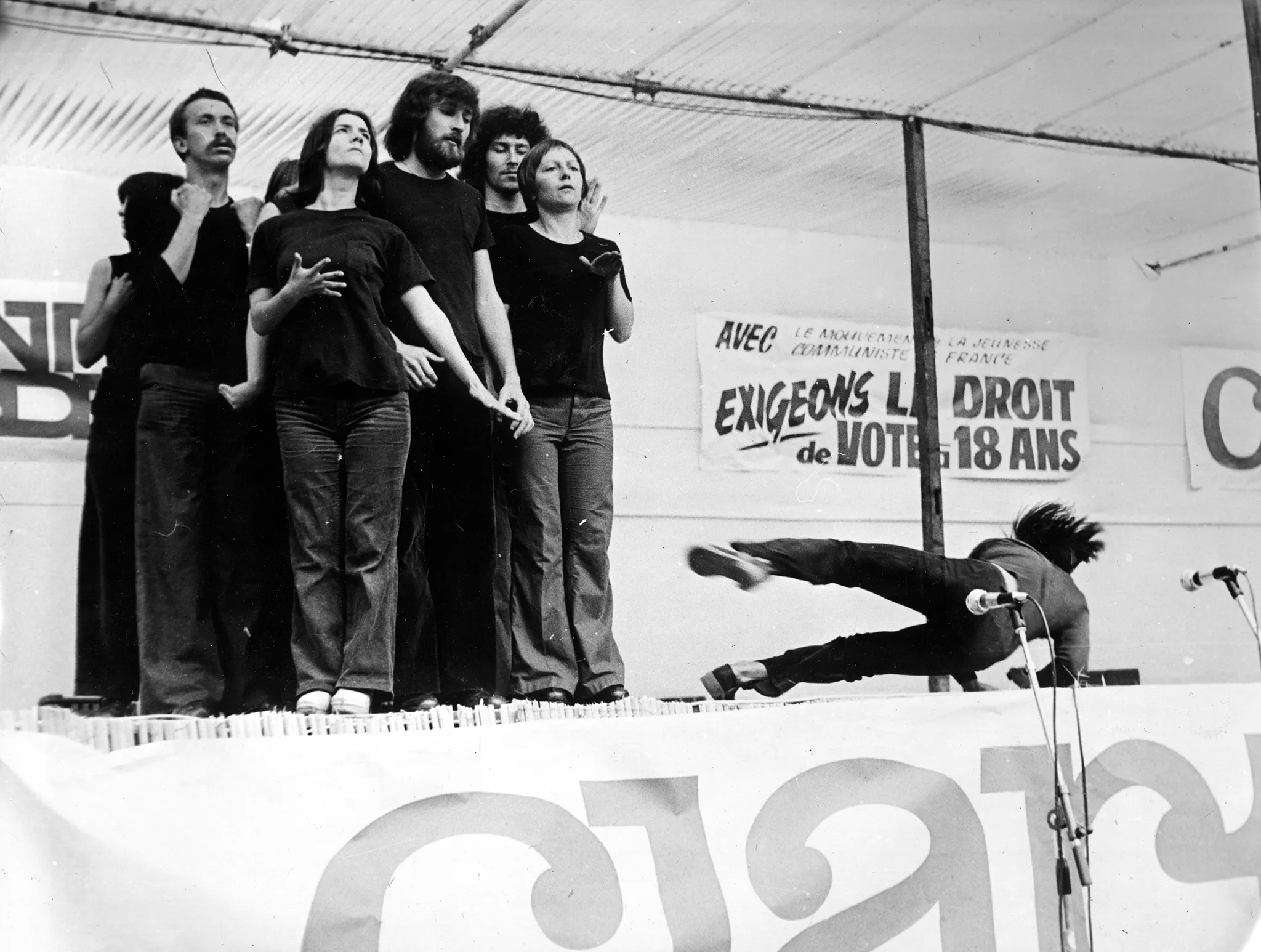 Grupa aktorów i aktorek na zadaszonej, plenerowej scenie. Grupa jest stłoczona. Aktorzy i aktorki ubrani są w jeansy i czarne podkoszulki. Po prawej stronie od grupy jeden aktor upada na scenę. Z tyłu sceny widać napisy po francusku. Pod sceną stoją mikrofony.