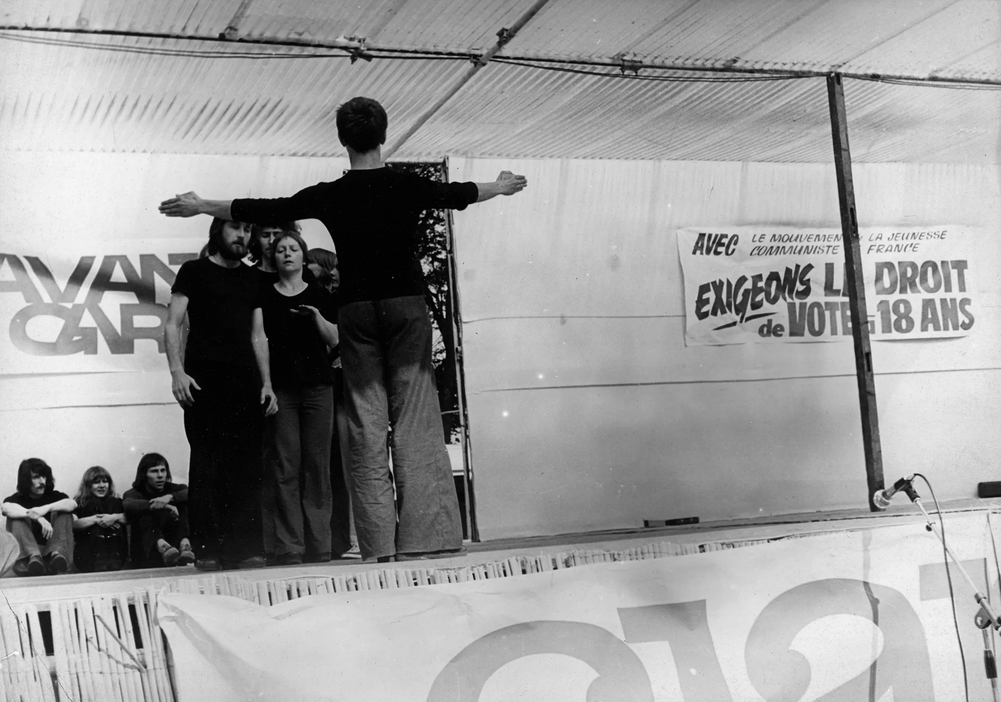 Grupa aktorów i aktorek na zadaszonej, plenerowej scenie. Przed nimi, stoi zwrócony do nas tyłem aktor wyglądający, jakby dyrygował grupie. Z tyłu sceny siedzi troje aktorów przyglądających się grupie. Aktorzy i aktorki ubrani są w jeansy i czarne podkoszulki. Z tyłu sceny widać napisy po francusku. Pod sceną stoją mikrofony.