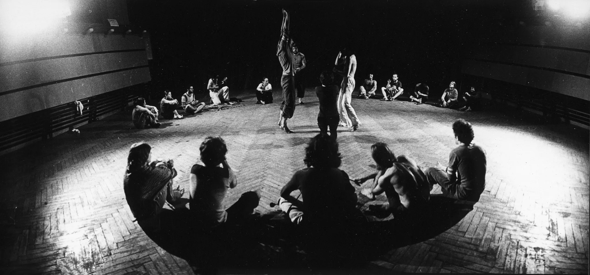 Aktorzy i aktorki siedzą na podłodze z panelu, tworząc duży okrąg. W środku okręgu czterech aktorów. Wyglądają, jakby tańczyli. Reszta aktorów im się przygląda.