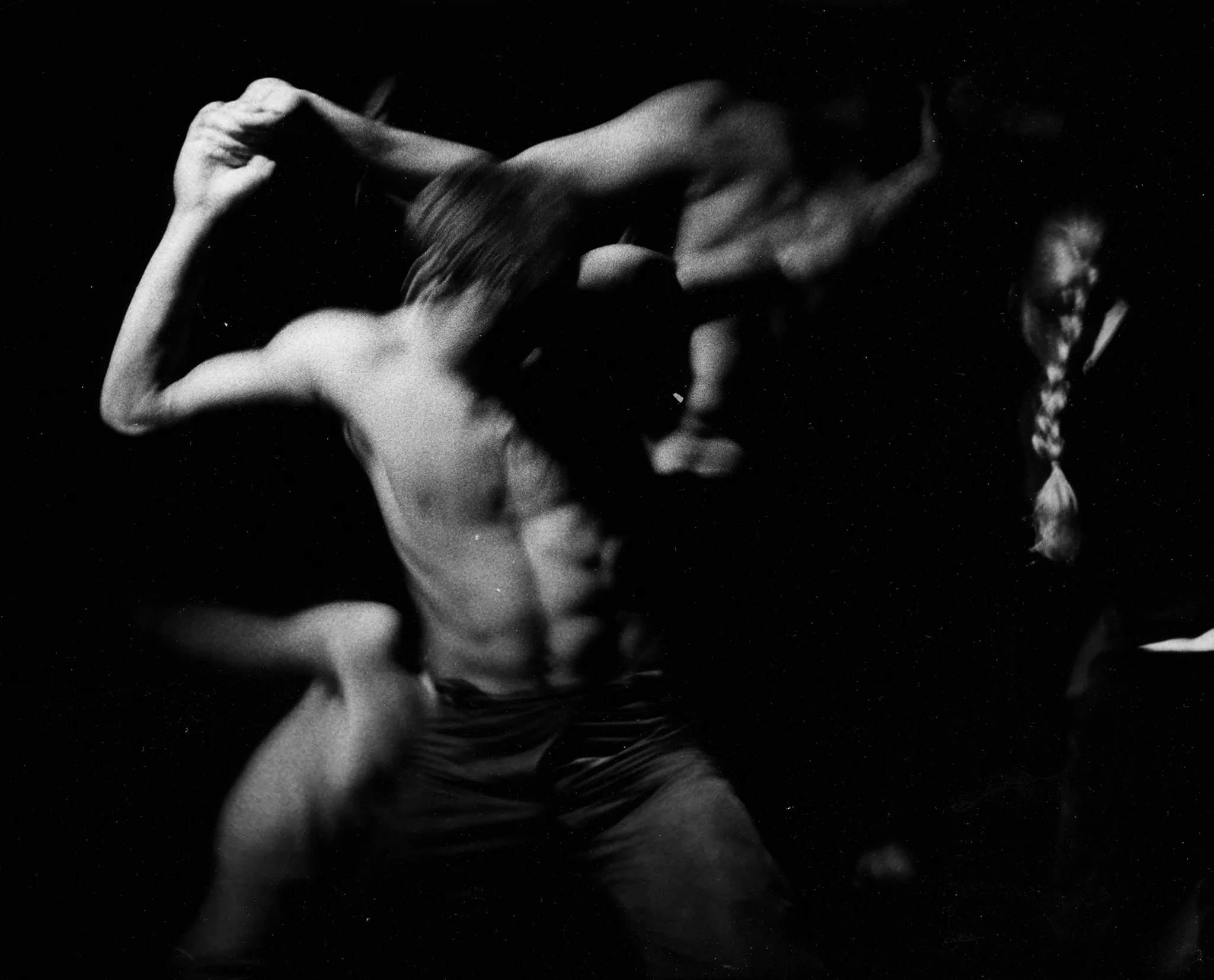 Niewyraźne, rozmazane zdjęcie. Aktor z wysportowanym nagim torsem trzyma kogoś na swoich barkach. Po prawej stronie stoi zwrócona do nas plecami aktorka. Ma blond włosy spięte w warkocz.