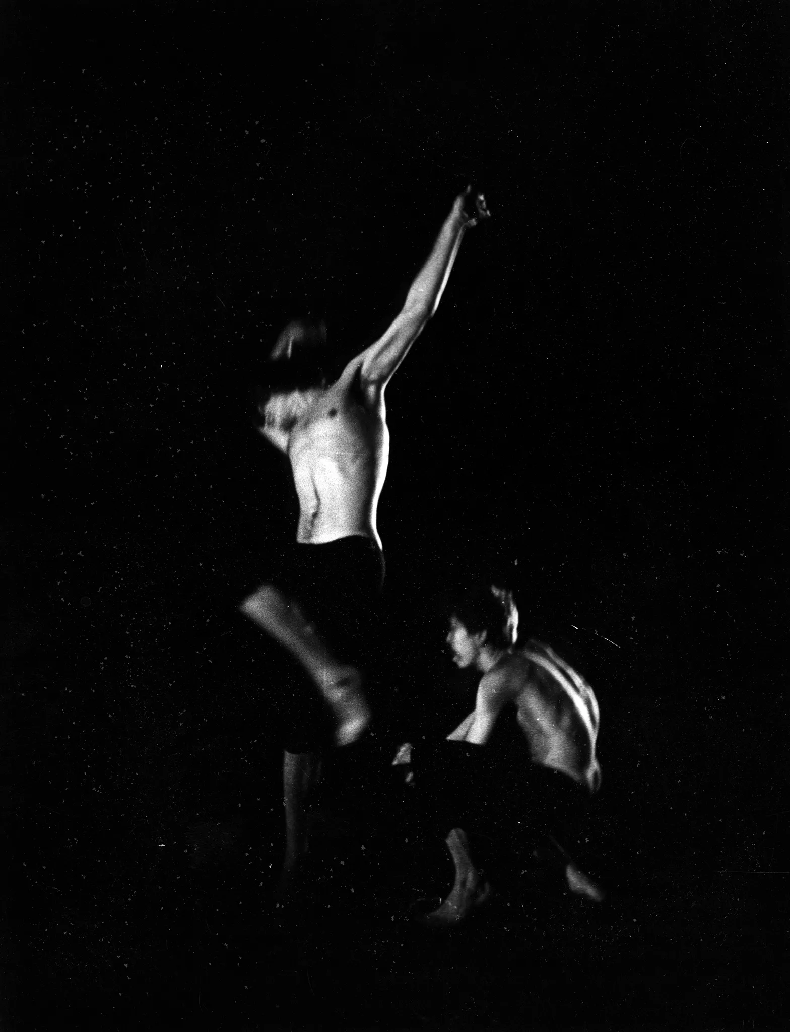 W centrum aktor uchwycony w skoku. Ma ręce wyciągnięte do góry. Ubrany jest w czarne spodenki, ma nagi tors. Nie widać jego głowy. Po jego prawej stronie klęczy aktor. Jest ubrany w czarne spodenki, ma nagi tors.