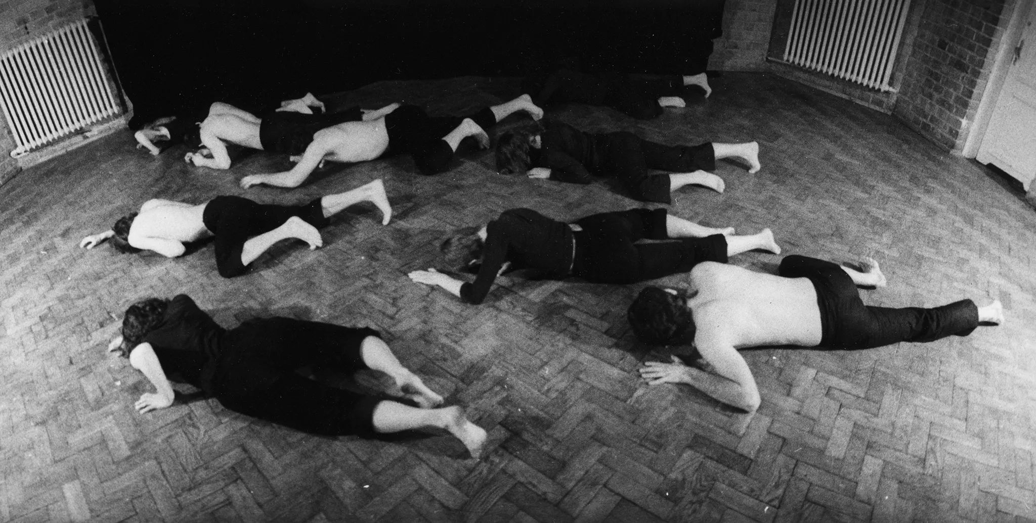 Grupa aktorów i aktorek tarza się po podłodze. Niektórzy są ubrani w czarne spodenki i czarne podkoszulki. Niektórzy nie mają podkoszulek. Są boso. Znajdują się na podłodze z paneli. W tle widać drzwi i kaloryfery.