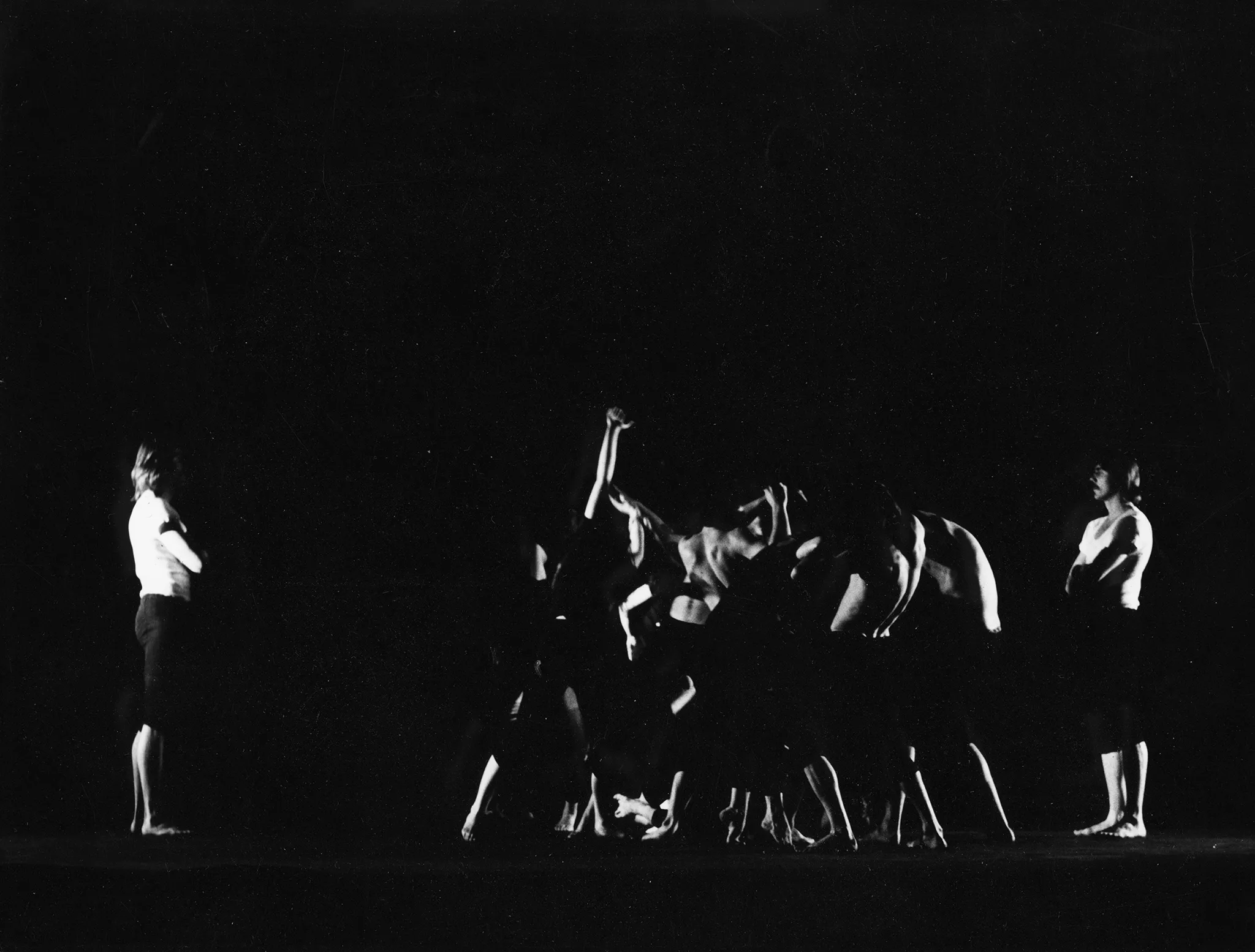 Na środku pustej sceny grupa aktorów wykonuje akrobacje. Są w bliskim kontakcie cielesnym. Po lewej i prawej stronie stoją zwróceni do nas profilem bardzo podobni do siebie aktorzy ubrani w czarne podkoszulki i czarne spodenki. Są boso. Patrzą na siebie. Grupa jest ustawiona bliżej aktora z prawej strony.
