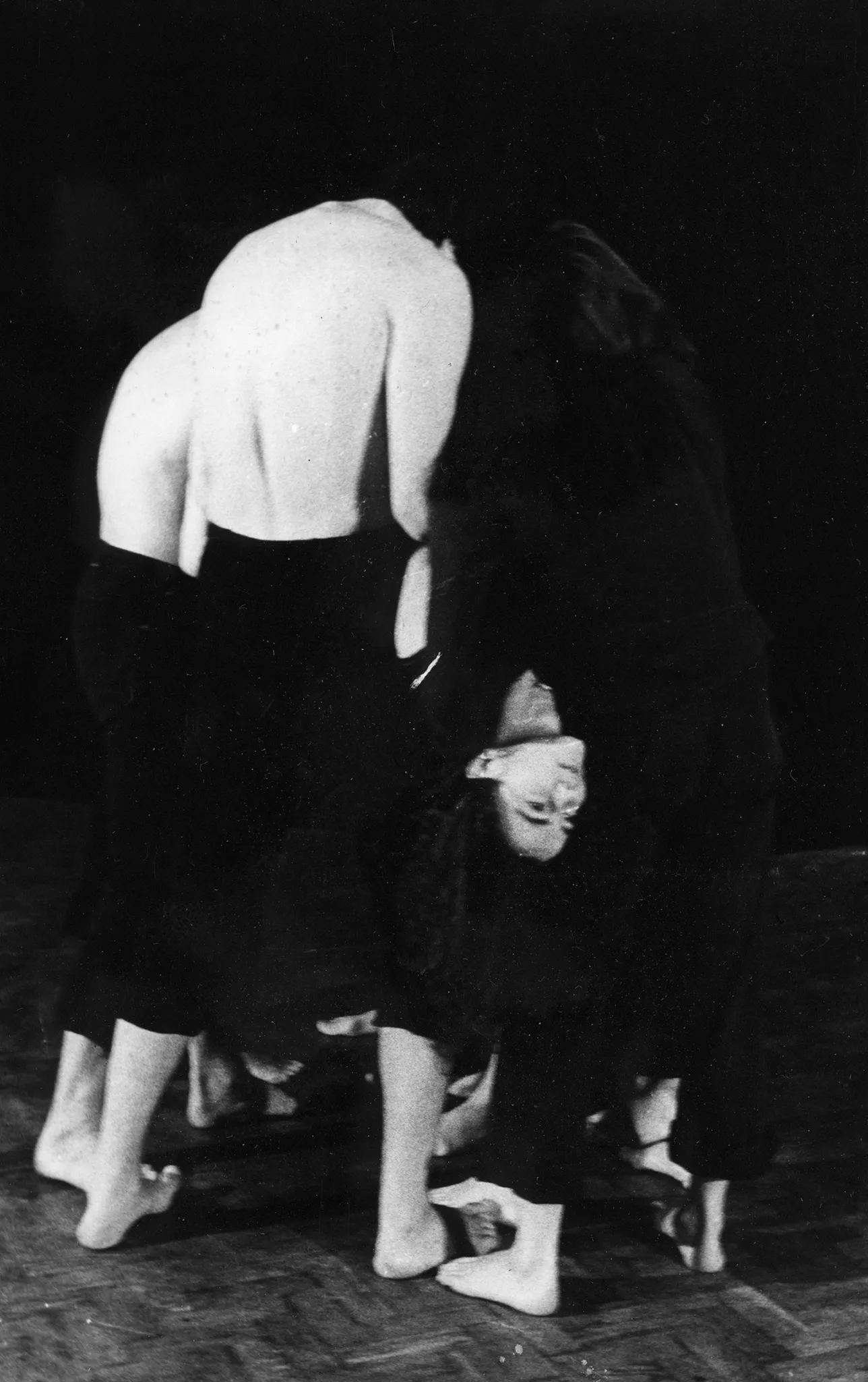 Grupa aktorów i aktorek ustawiona w okręgu. Aktorzy mają na sobie czarne spodenki, ich torsy są nagie. Aktorki ubrane są w czarne spodenki i czarne podkoszulki. Grupa stojąca w okręgu trzyma na rękach aktorkę leżącą brzuchem do góry. Aktorka patrzy przed siebie, jej włosy zwisają.