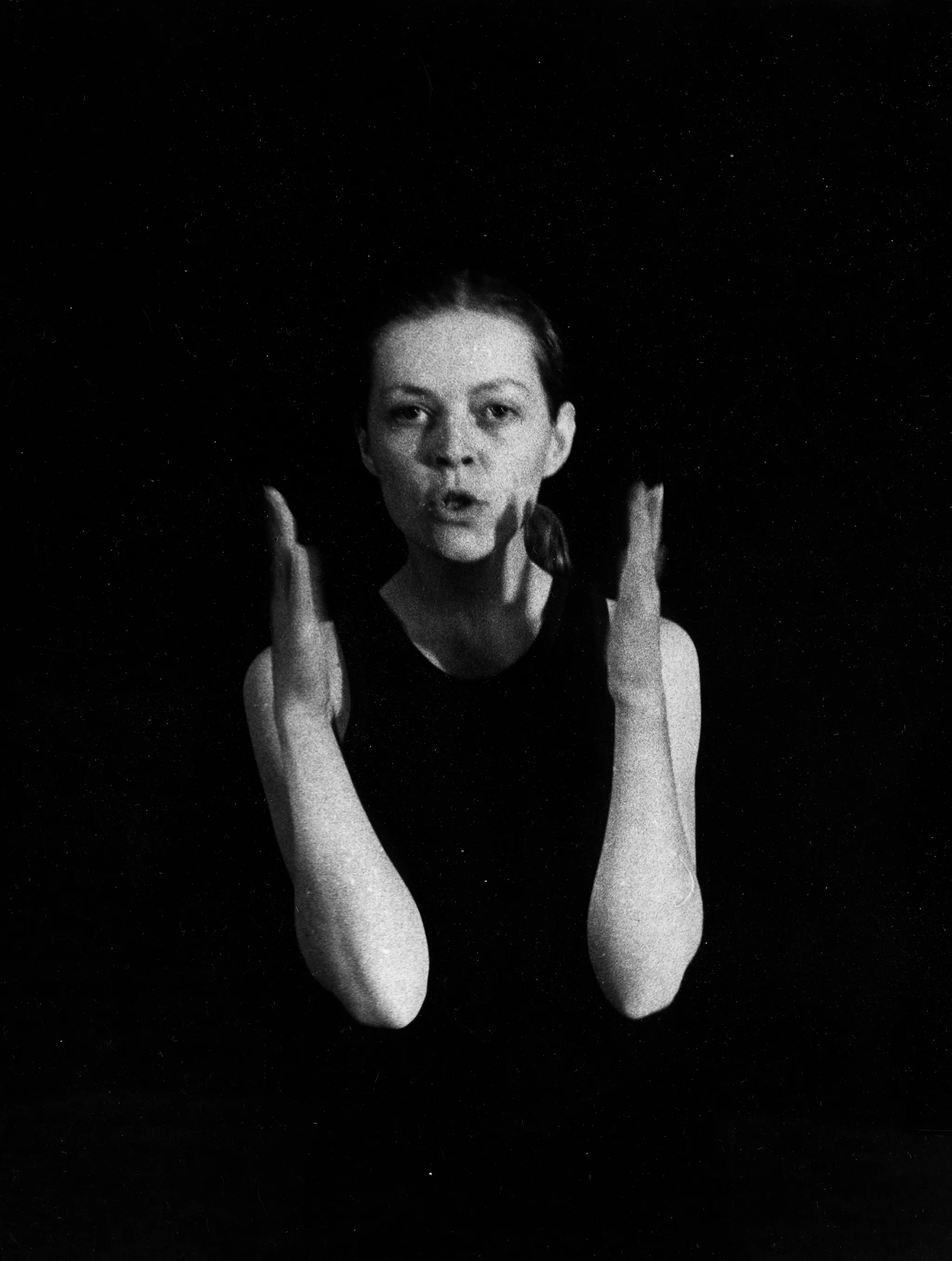 Zwrócona do nas twarzą aktorka. Ma zgięte, wyciągnięte przed siebie ręce. Wygląda, jakby coś mówiła. Ubrana jest w czarną koszulkę na ramiączka.