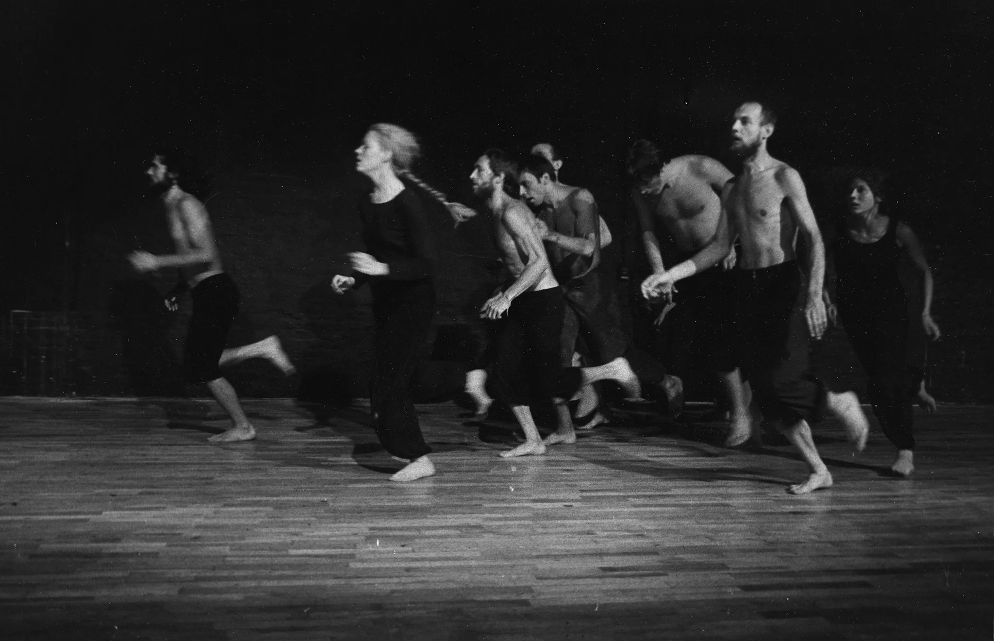Grupa aktorów i aktorek biegnie po panelowej podłodze. Aktorzy ubrani są w czarne spodenki, mają nagie torsy. Aktorki ubrane są w czarne spodenki i czarne podkoszulki. Wszyscy są boso.