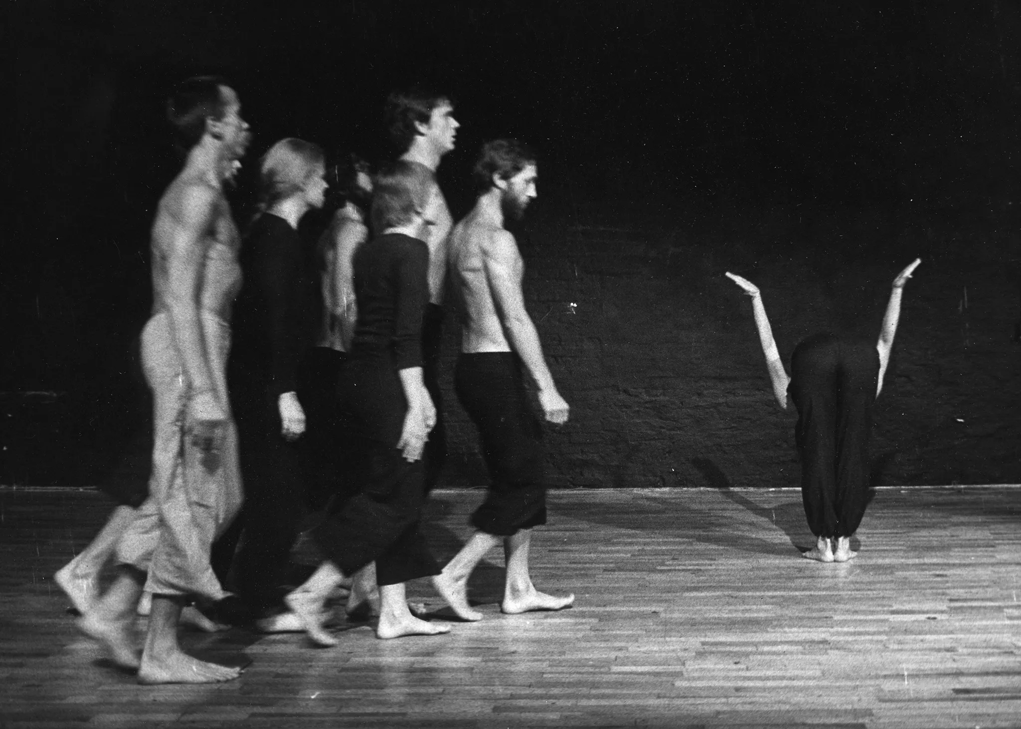 Od lewej strony idzie zwrócona profilem do widza grupa aktorów i aktorek. Aktorzy mają nagie torsy i czarne spodnie poza jednym aktorem, którego spodnie są jasne. Aktorki ubrane są w czarne spodnie i czarne koszulki. Wszyscy są boso. Po prawej stronie stoi odwrócona plecami w stronę widza aktorka. Jest pochylona do przodu i trzyma ręcę uniesione po bokach. Jest boso. Podłoga, na której stoją aktorzy, wygląda na drewnianą.