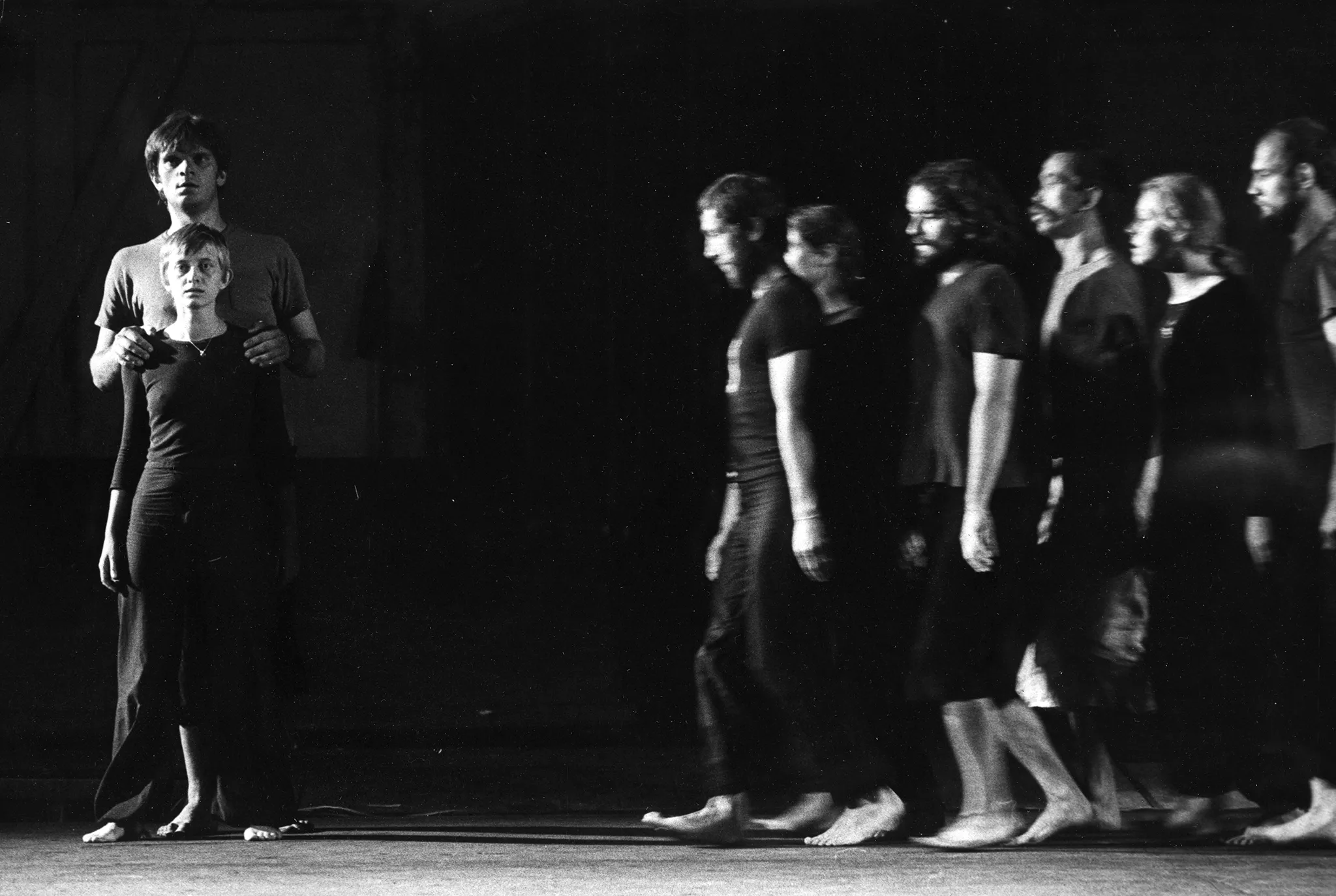 Po prawej stronie grupa aktorów i aktorek zwrócona do nas profilem w szeregu idzie w lewą stronę. Są ubrani w czarne spodenki i czarne podkoszulki. Są boso. Po lewej stronie stoi zwrócona twarzą w nasza stronę aktorka. Tuż za nią stoi zwrócony twarzą do nas wyższy od niej o głowę aktor obejmujący jej ramiona. Oboję są ubrani w czarne spodnie i ciemne podkoszulki. Są boso.