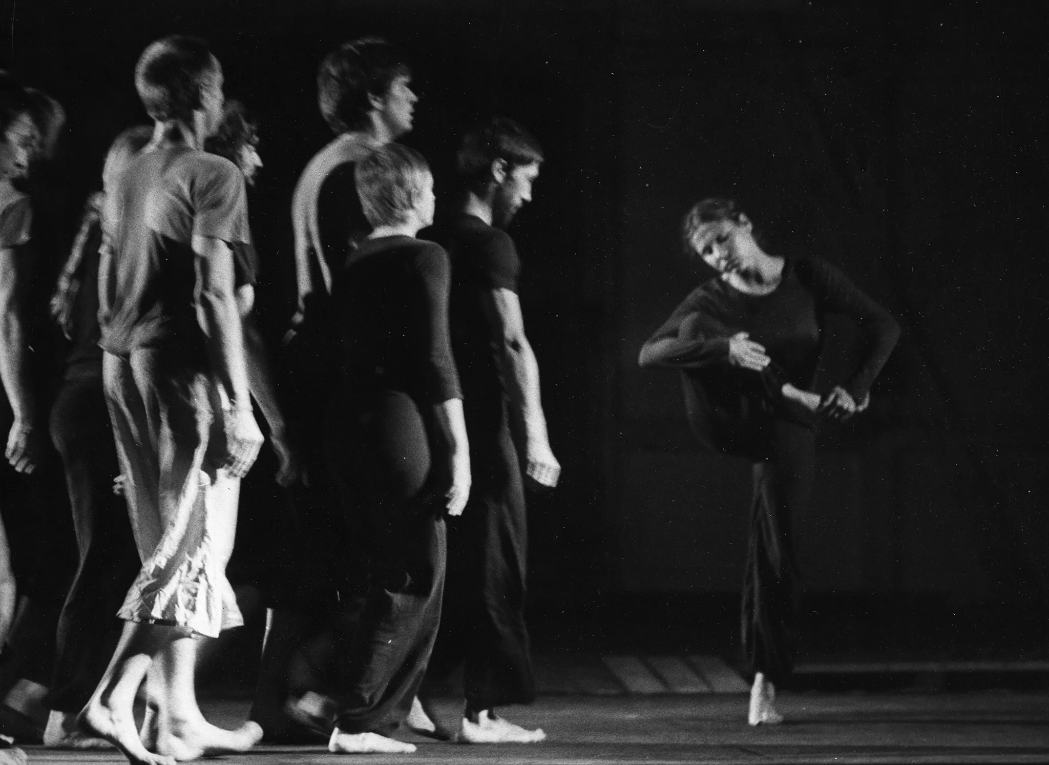 Od lewej stronie idzie zwrócona profilem do widza grupa aktorów i aktorek. Ubrani są w ciemne spodnie i koszulki z wyjątkiem jednego aktora, którego spodnie są jasne. Wszyscy są boso. Po prawej stronie stoi aktorka. Ubrana jest w czarne spodnie i czarną koszulkę z długim rękawem. Stoi na jednej nodze. Drugą nogę ma zgiętą, trzyma ją rękami w górze. Patrzy w dół.