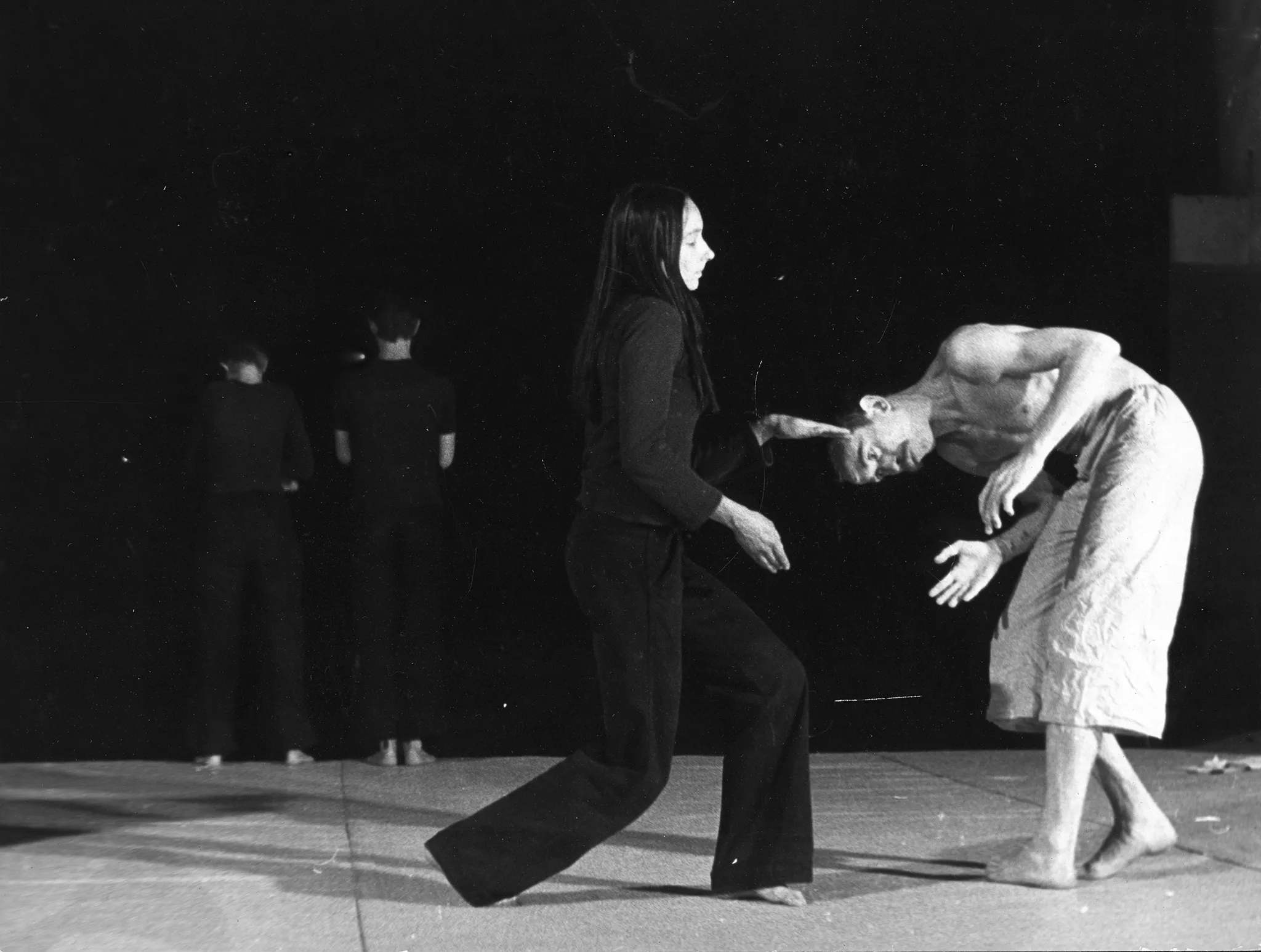 W centrum pustej, jasnej sceny stoi aktorka zwrócona do widza profilem. Jest ubrana w czarne spodnie i czarną koszulkę z długim rękawem. Jest boso. Jej włosy są długie i czarne. Ma ugięte obie nogi - jedną z przodu, drugą z tyłu. Gestykuluje dłońmi. Obok niej stoi mocno zgarbiony aktor. Ma jasne spodenki i nagi tors. Jest boso. Patrzy w podłogę. Po lewej stronie, w głębi stoi dwoje aktorów odwróconych do widzów plecami. Są ubrani na czarno.