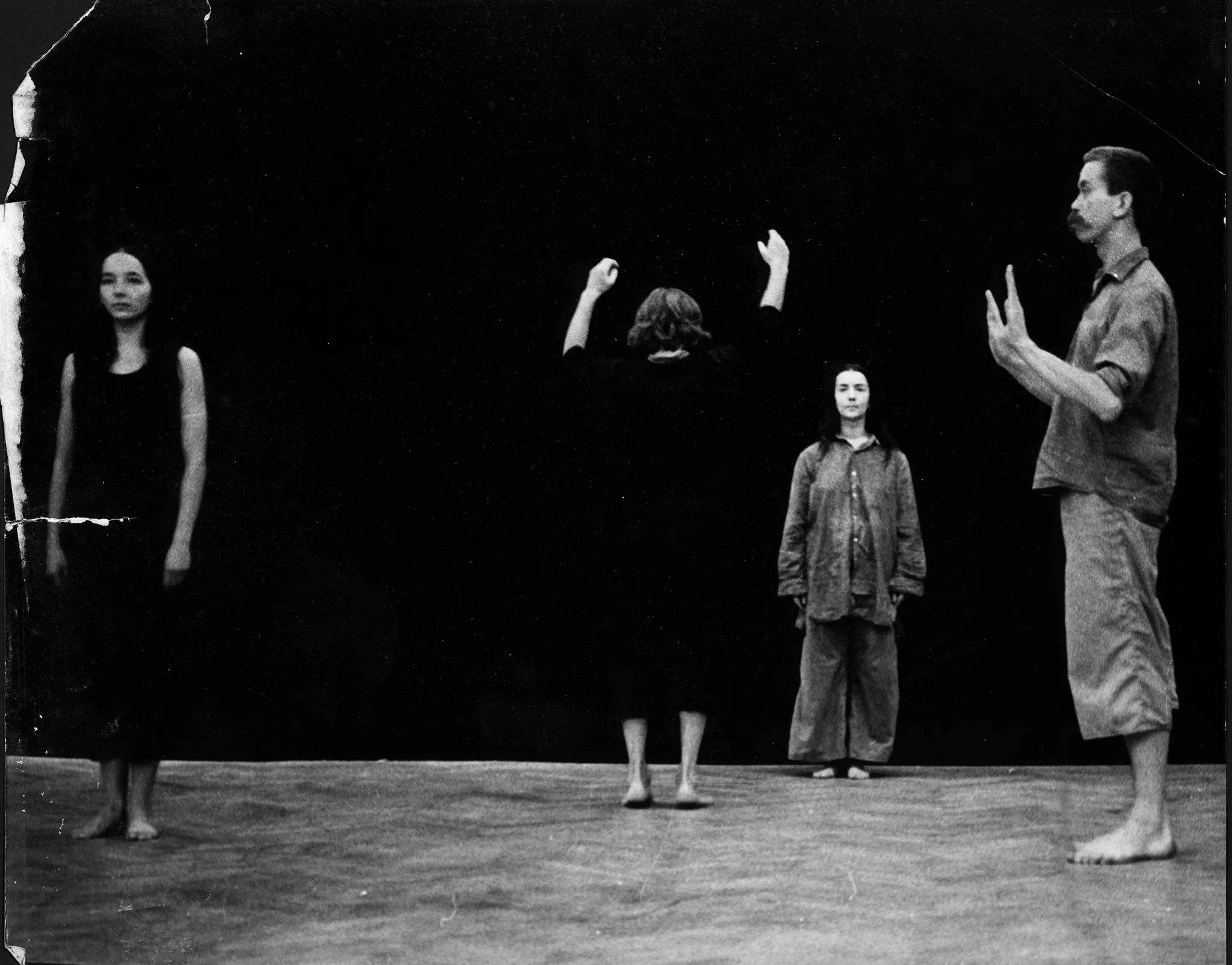 Po lewej stronie, zwrócona twarzą do widzów stoi aktorka. Ubrana jest na czarno, jest boso. W centrum sceny naprzeciwko siebie stoją dwie aktorki. Jedna, bliżej widzów, jest zwrócona do nich plecami. Ma uniesione ręce. Aktorka stojąca w głębi sceny zwrócona jest do widzów twarzą. Aktorki mają ciemne ubrania. Są boso. Patrzą na siebie nawzajem. Po prawej stronie, zwrócony profilem do widzów stoi aktor. Ma obie ręce wyciągnięte przed siebie. Ubrany jest w jasne spodenki i ciemną koszulę. Jest boso.