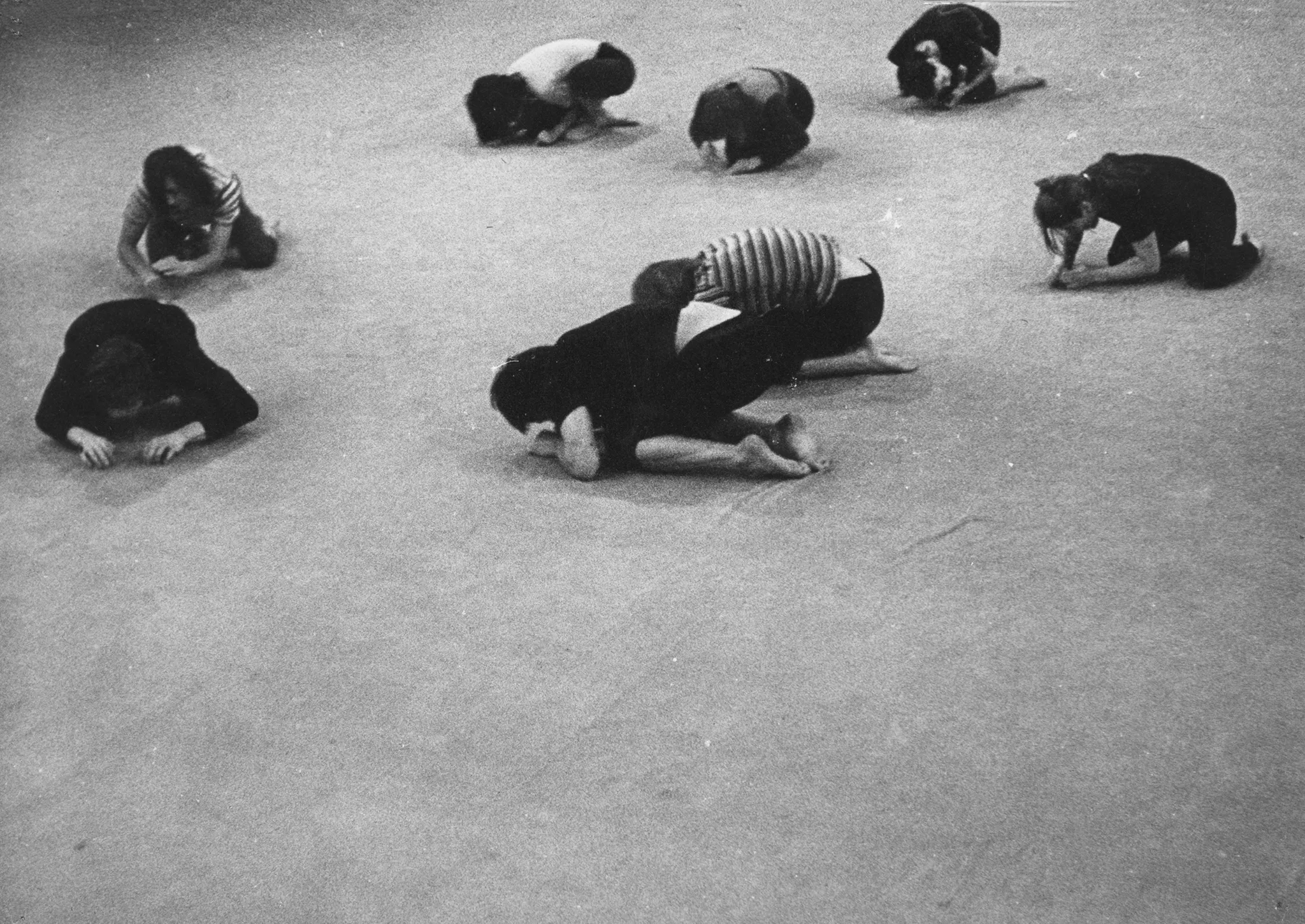 Grupa aktorów i aktorek leży skulona na podłodze. Wszyscy leżą od siebie w pewnej odległości. Są boso. Niektórzy ubrani są w czarne spodenki i czarne podkoszulki. Niektórzy mają podkoszulki w czarno-białe paski.