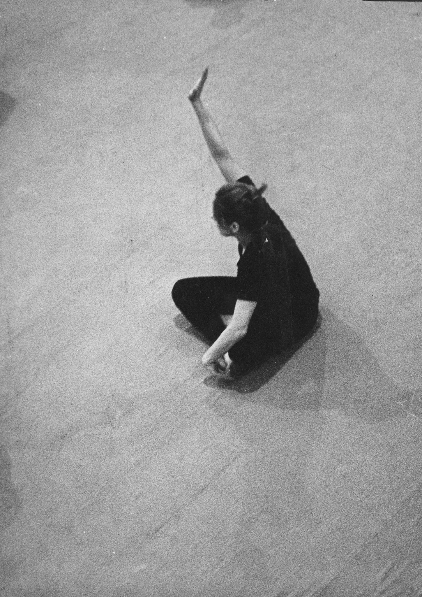 Aktorka siedzi w siadzie skrzyżnym. Widać ją od góry. Aktorka unosi jedną rękę. Ubrana jest w czarne spodnie i czarną podkoszulkę.