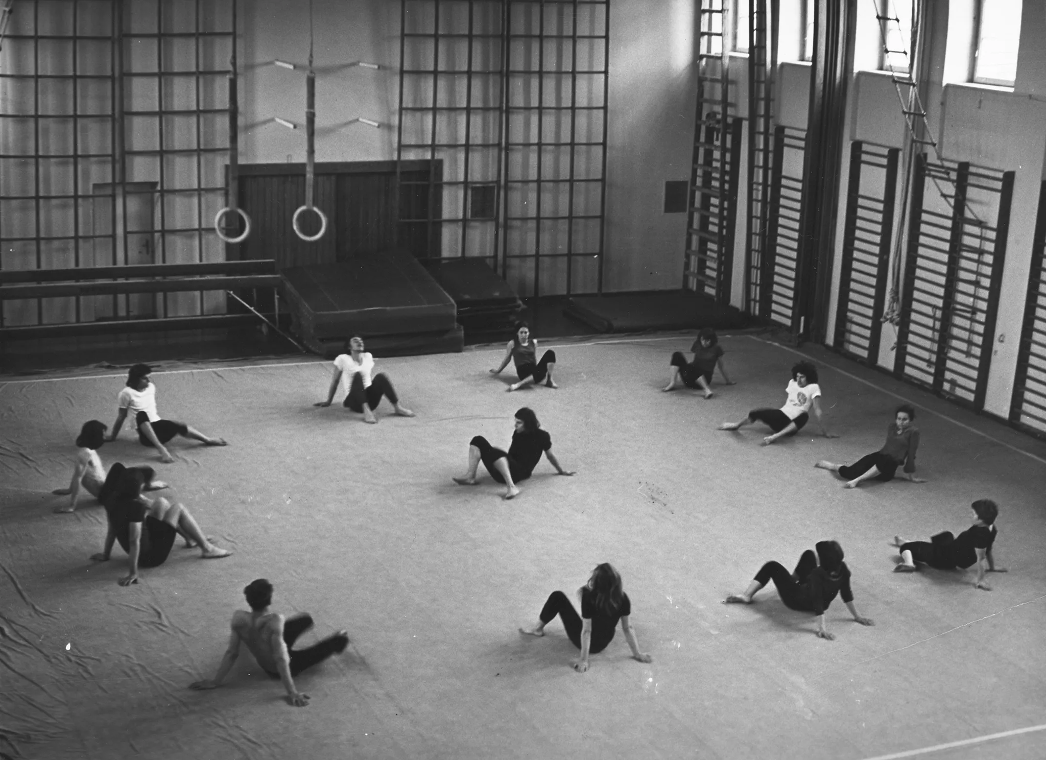 Grupa aktorów i aktorek na sali gimnastycznej. Przy ścianach stoją drabinki, z sufitu zwisają liny gimnastyczne. Aktorzy i aktorki siedzą na podłodze w okręgu. Są zgarbieni i mają wyprostowane nogi. Rozciągają się.