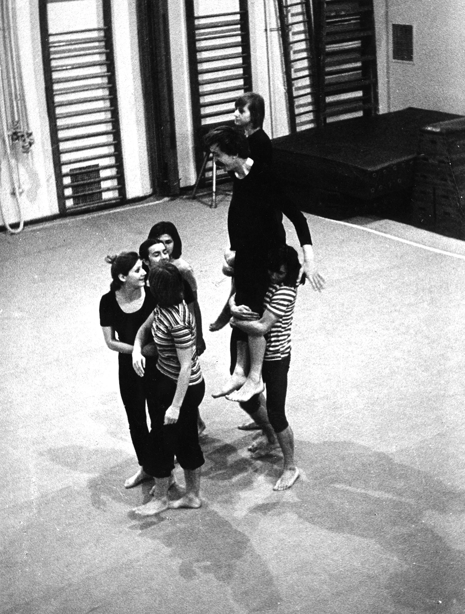 Grupa aktorów i aktorek na sali gimnastycznej. W tle drabinki gimnastyczne i duży materac. Aktorzy i aktorki są boso. Niektórzy ubrani są w czarne spodenki i czarne podkoszulki, inni - w czarno-białe podkoszulki w paski. Jedna z aktorek podnosi aktora na rękach. Wszyscy są uśmiechnięci.