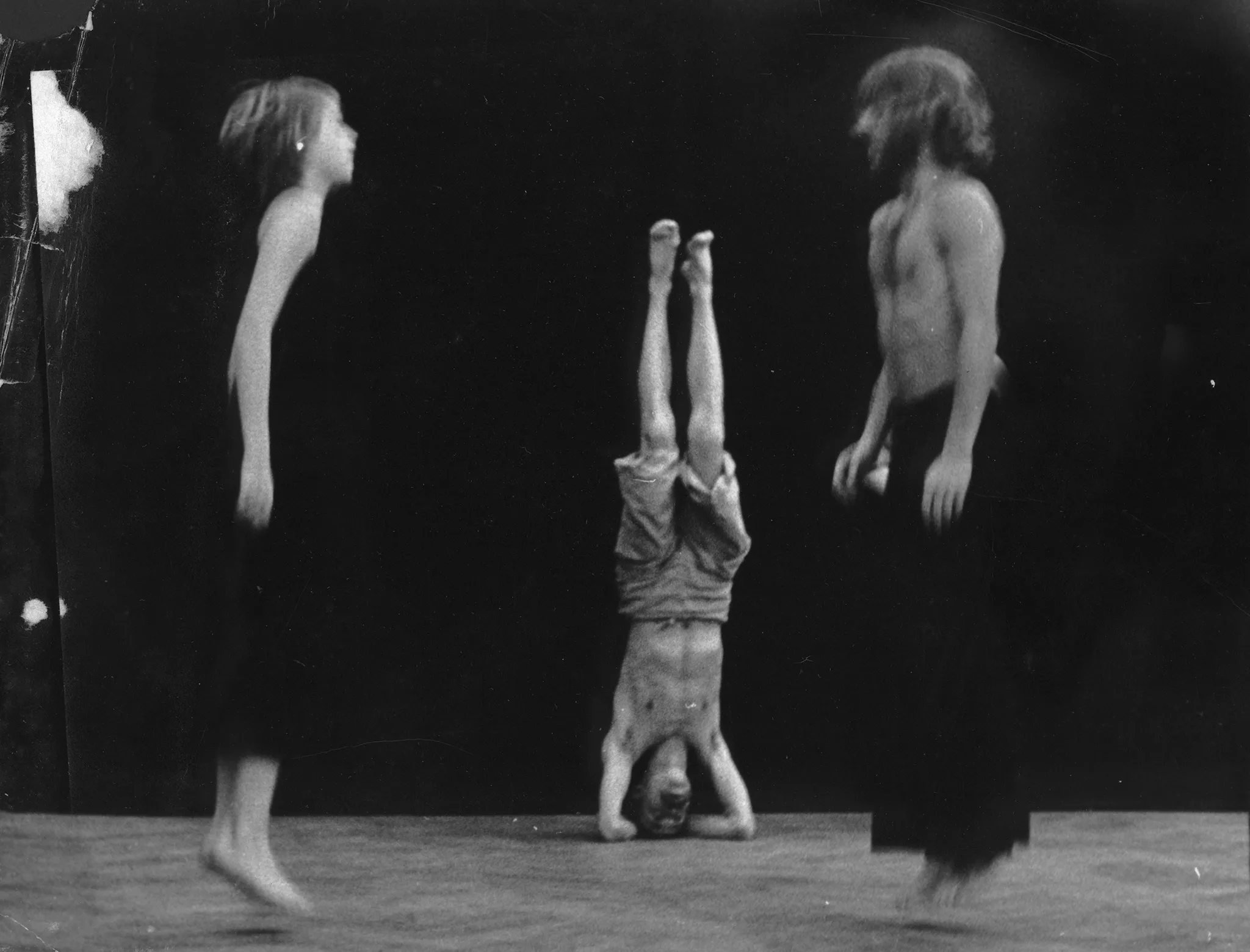Aktorka i aktor zwróceni profilem do widza, twarzami do siebie. Aktorka po lewej stronie, aktor - po prawej. Aktorka ubrana jest na czarno, aktor ubrany jest w czarne spodnie i ma nagi tors. Oboje są boso. Skaczą. W centrum, w oddali aktor stoi na głowie. Ma nagi tors, jego spodenki opadają.
