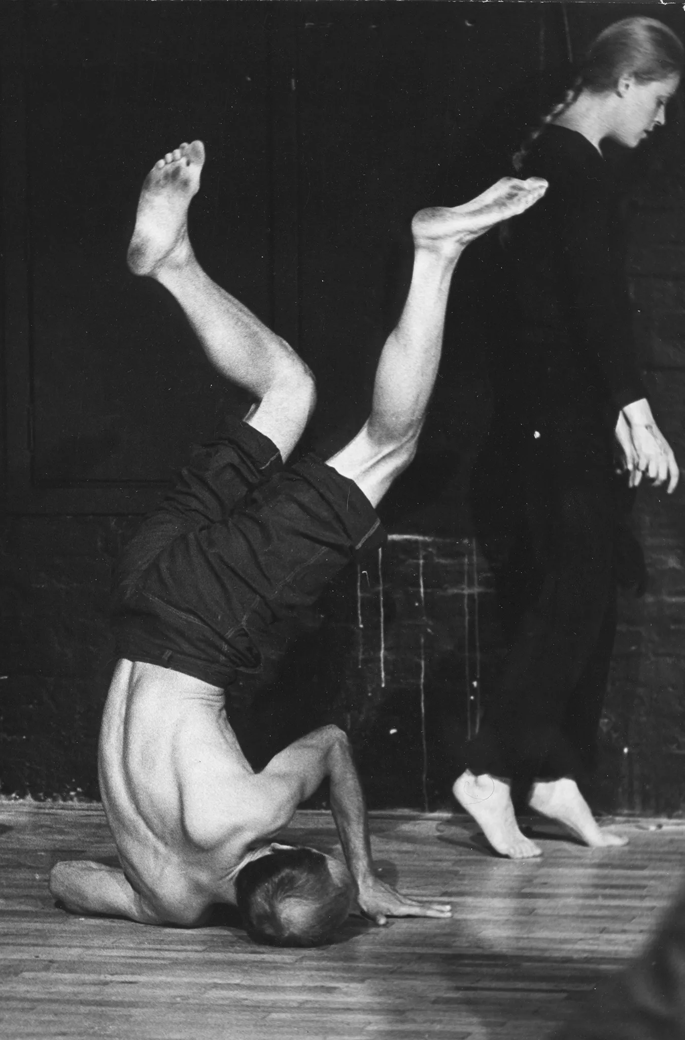 Aktor stoi na przekrzywionej głowie podpierając się rękami. Jego ciało jest wygięte. Ustawiony jest plecami w stronę widza. Po prawej stronie aktorka stoi na palcach. Jest ubrana na czarno i jest boso. Jej włosy są splecione w warkocz.