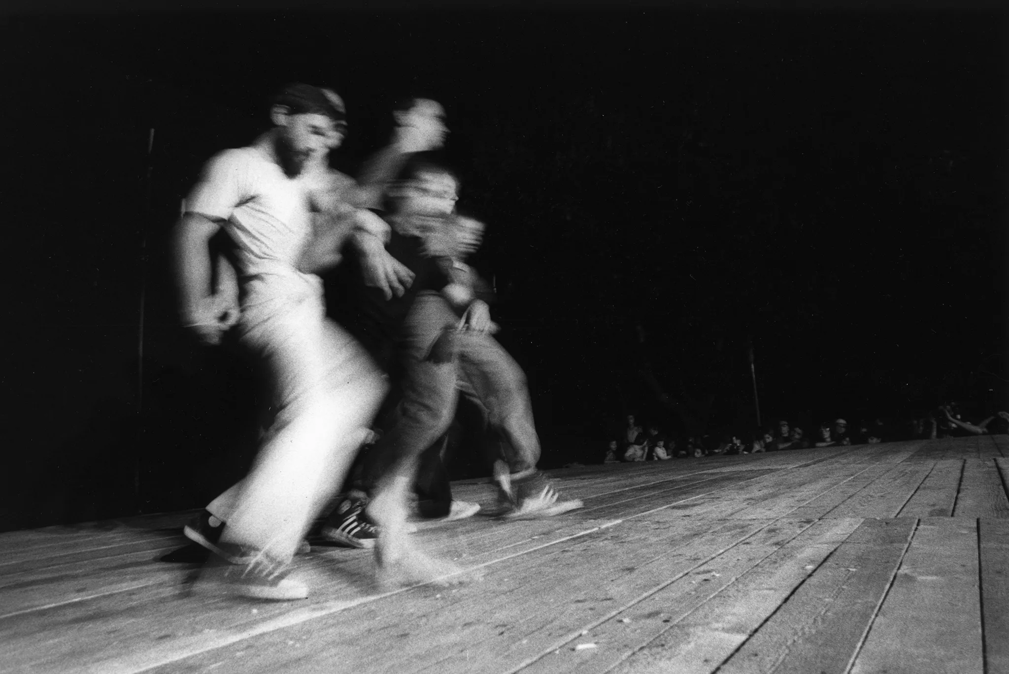 Niewyraźne, rozmazane zdjęcie. Od lewej strony idzie grupa aktorów. Są bardzo blisko siebie, idą na ugiętych nogach. Znajdują się na drewnianej podłodze. W tle widać widownię.