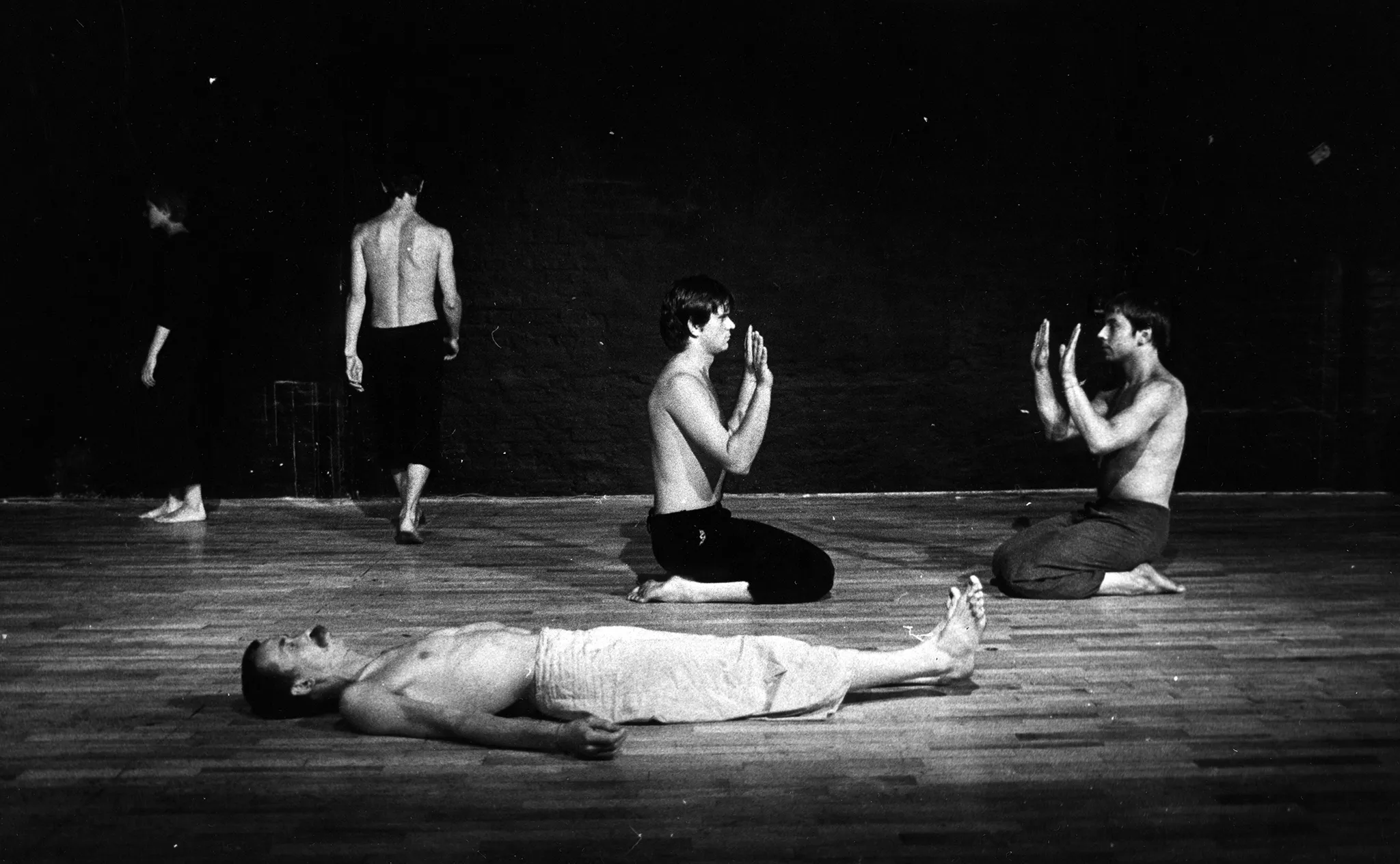 Po lewej stronie stoi zwrócona profilem do widza aktorka. Ubrana jest w czarne spodenki i czarną podkoszulkę. Jest boso. Obok niej, stoi zwrócony tyłem do widza aktor. Ubrany jest w czarne spodenki, ma nagi tors. Jest boso. Idzie w stronę tyłu sceny. W centrum, na pierwszym planie na plecach leży aktor. Ubrany jest w jasne spodenki, ma nagi tors. Patrzy w sufit i wyciągata przed siebie jedną rękę. Za nim klęczy dwóch aktorów. Ubrani są w ciemne spodenki, mają nagie torsy. Klęczą naprzeciwko siebie i patrzą na siebie nawzajem. Pokazują sobie wnętrza dłoni.