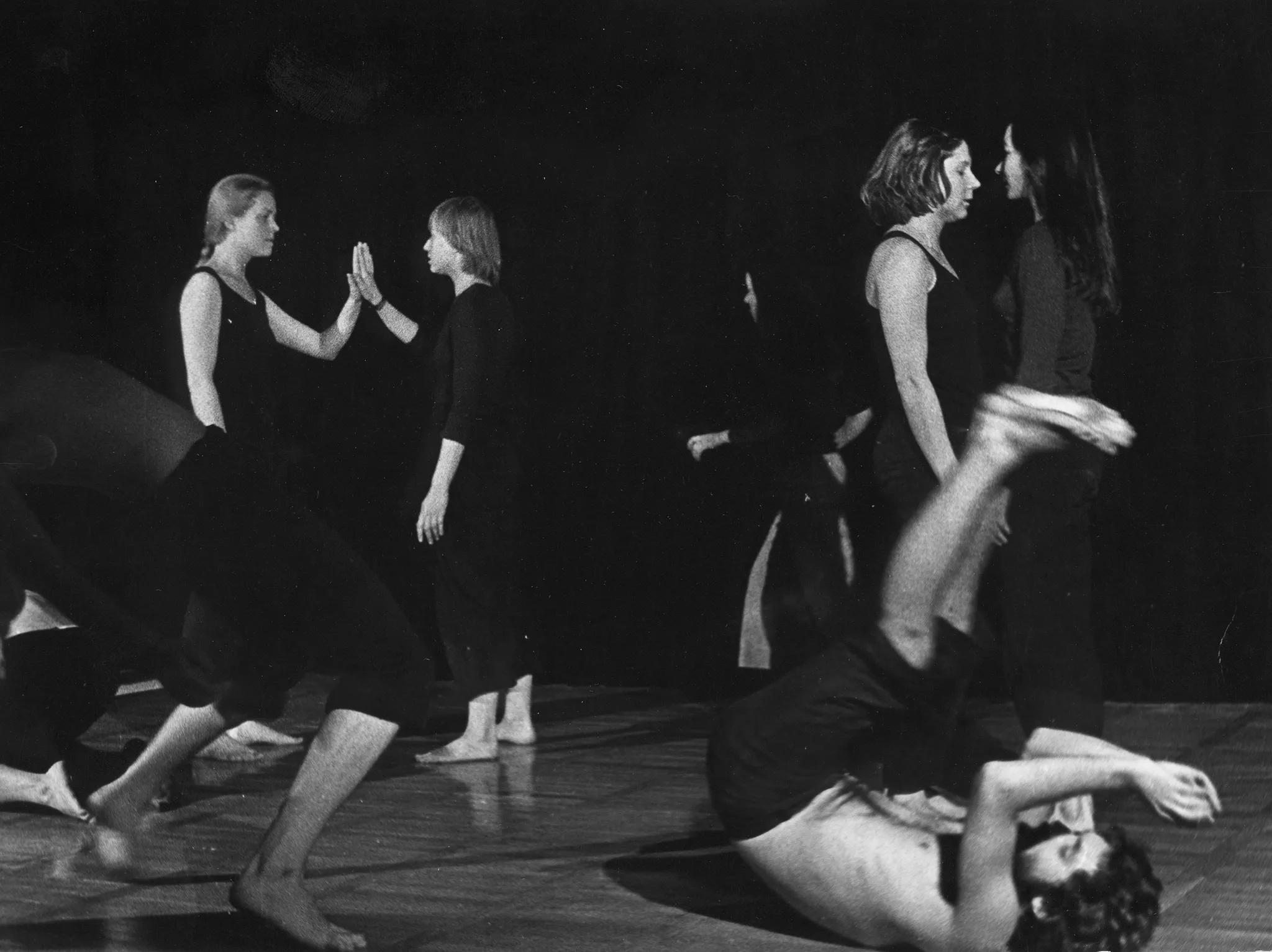 Grupa aktorów i aktorek. Na pierwszym planie dwóch aktorów wykonuje akrobatyczne ruchy na podłodze. Za nimi, w dwóch parach stoją cztery aktorki. W każdej parze aktorki są zwrócone do siebie twarzami. W oddali przechodzi przez scenę słabo widoczny aktor. Aktorzy ubrani są w czarne spodenki, mają nagie torsy. Aktorki ubrane są w czarne spodnie i czarne koszulki. Wszyscy są boso.