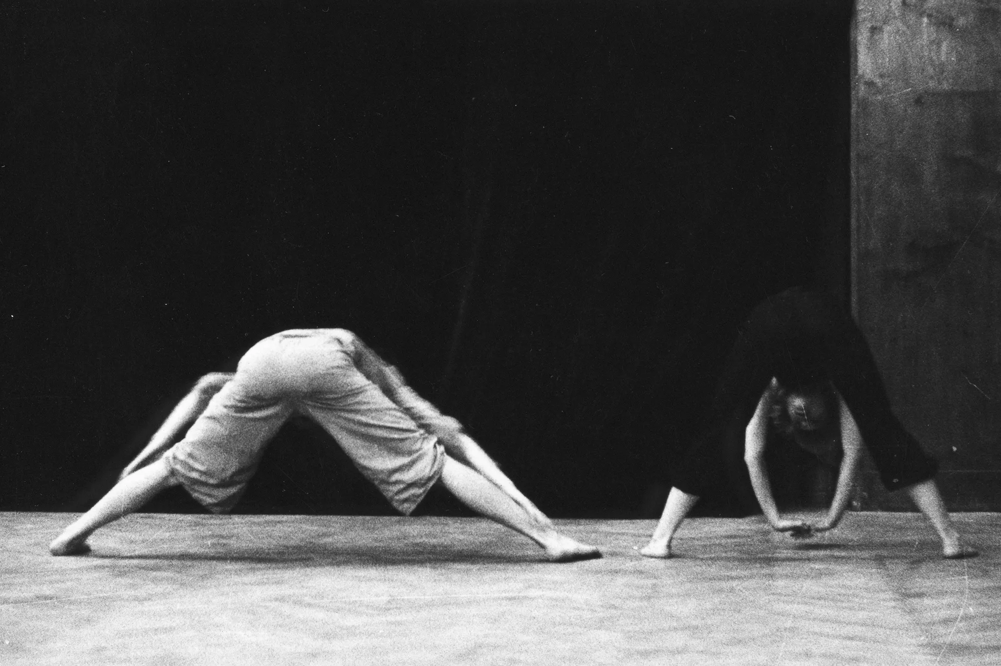 Aktor i aktorka stoją obok siebie. Aktor stoi po lewej stronie, aktorka po prawej. Mają nogi szeroko rozstawione i rozciągają się schylając głowę w stronę podłogi. Aktor ubrany jest w jasne ubrania, aktorka w czarne. Oboje są boso.